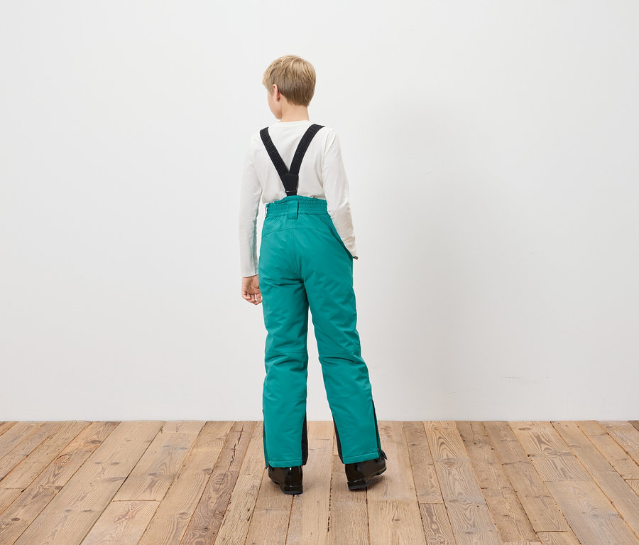 Egy fiú háttal áll a kamerának, türkiz síkabátot visel fekete nadrágtartóval.