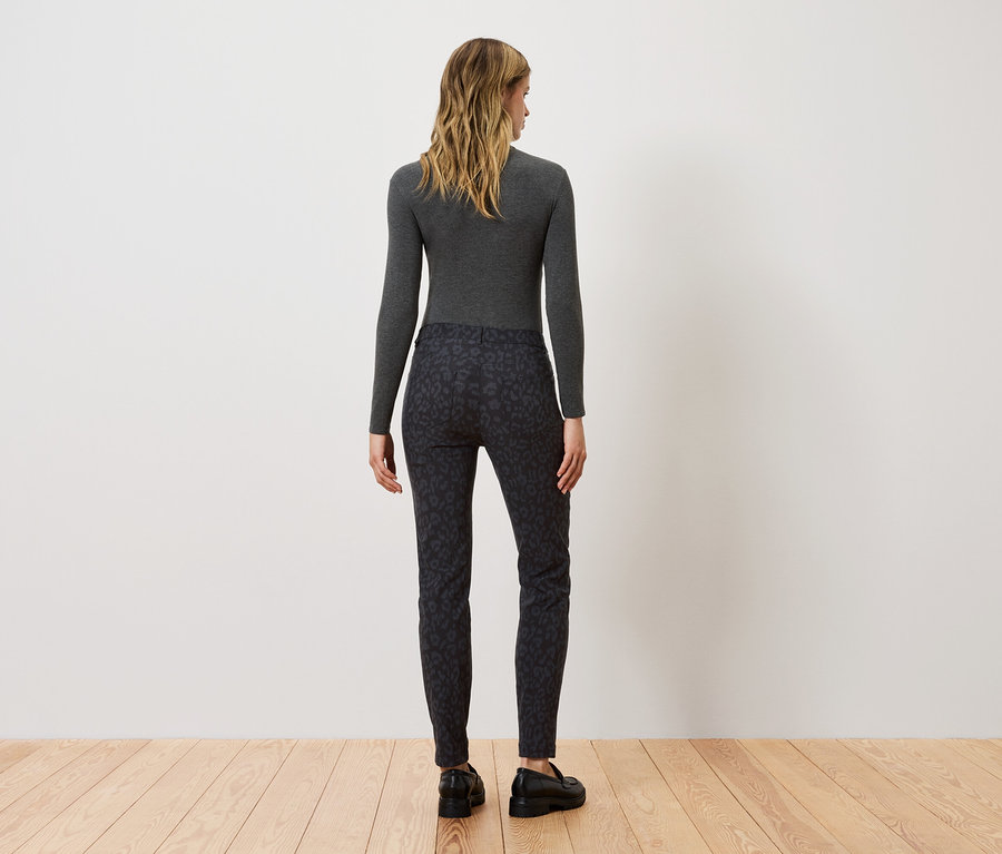 Egy nő háttal áll a kamerának, szürke hosszú ujjú felsőt és fekete mintás nadrágot visel.