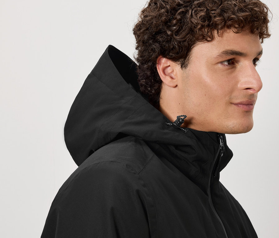 Egy barna, göndör hajú férfi fekete kapucnis kabátot visel.