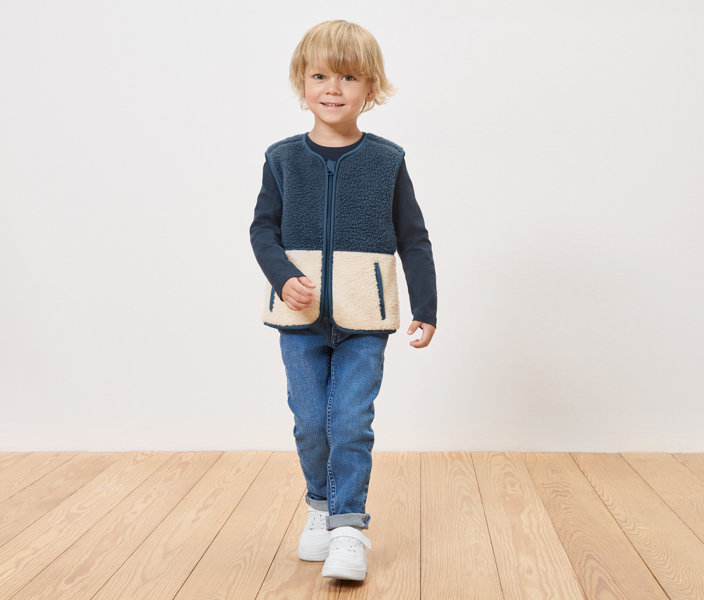 Egy fiú tengerészkék színű, cipzáras, krémszínű alsó betétekkel ellátott polár mellényben áll egy fapadlón.