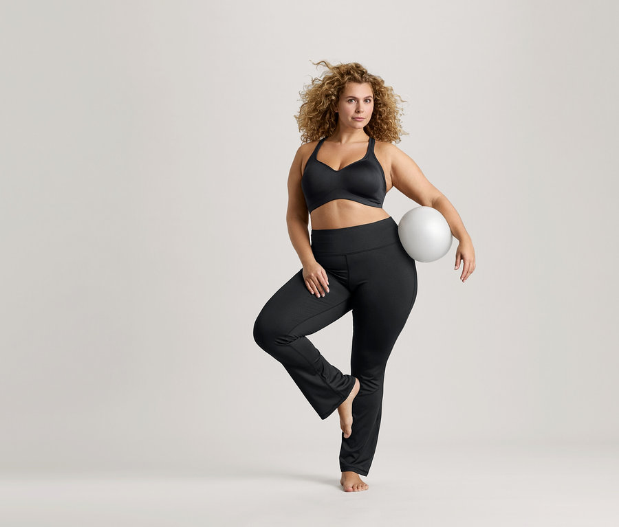 Göndör hajú nő egy lábon egyensúlyoz, fehér labdát tart, fekete sportmelltartót és fekete leggingset visel.