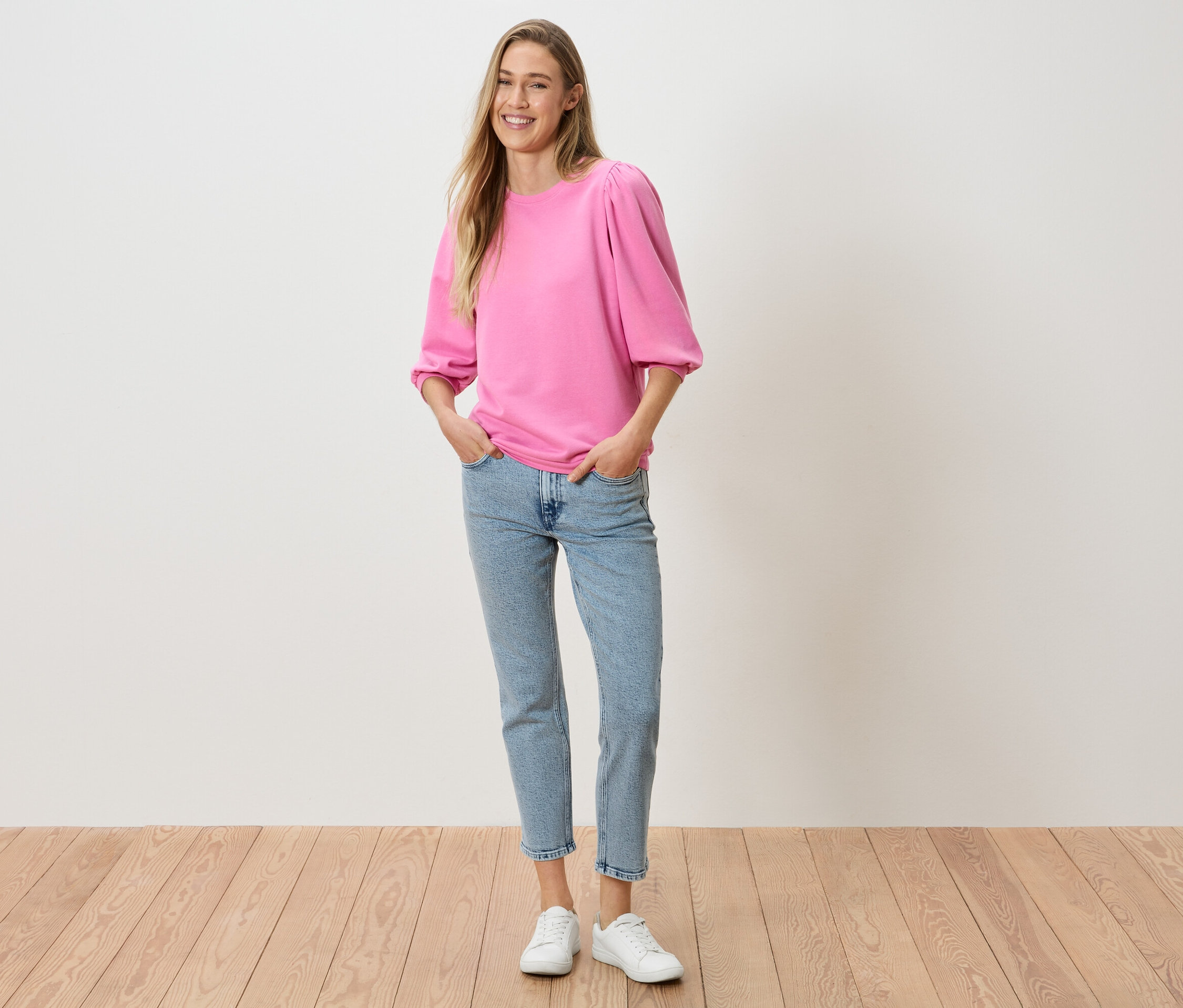 Egy nő a zsebében tartja a kezét, rózsaszín puffos ujjú felsőt, farmert és fehér tornacipőt visel.