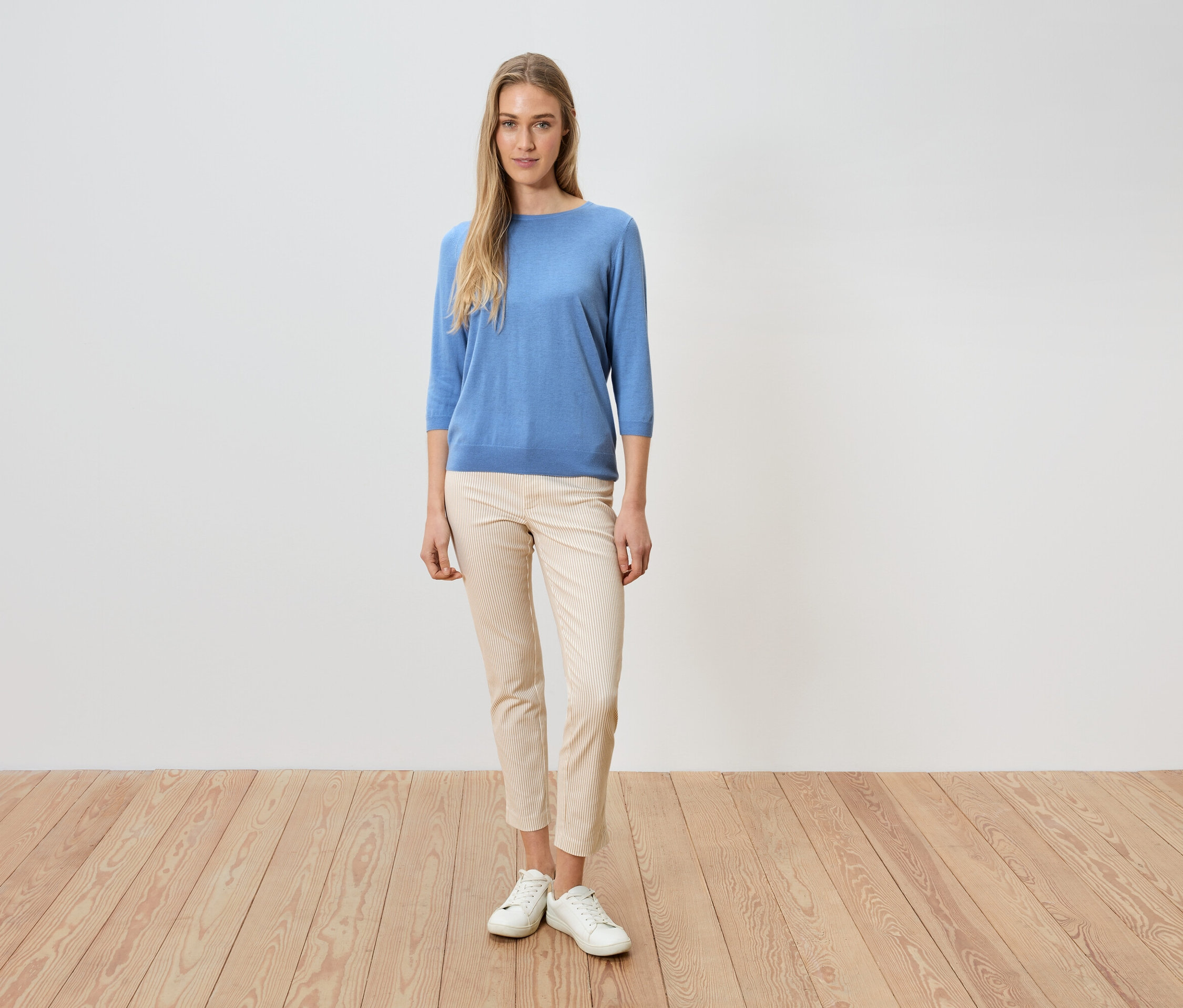 Egy nő áll egy fehér fal előtt, kék pulóvert, világos nadrágot és fehér cipőt visel.