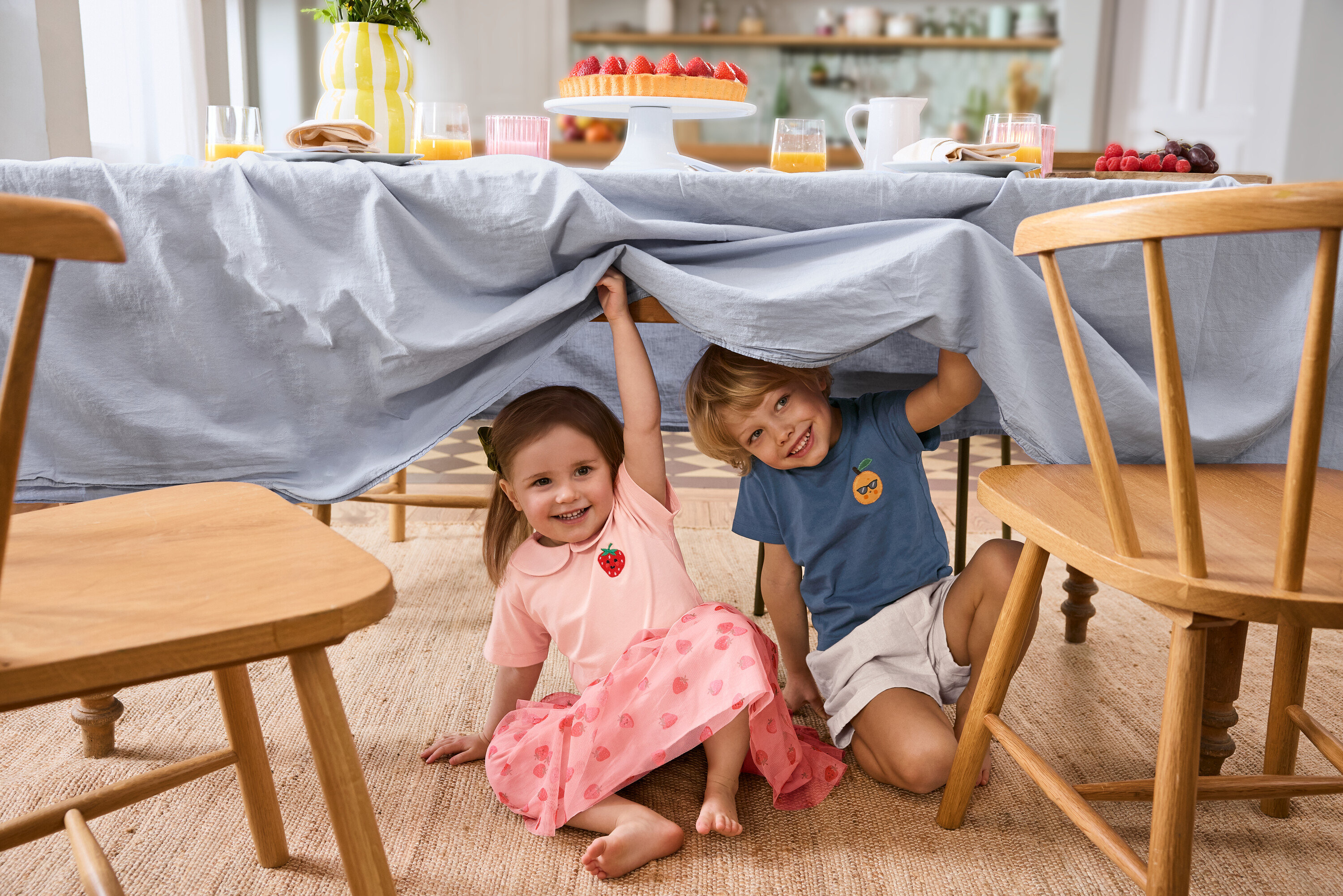 Két gyerek ül egy világoskék terítővel letakart asztal alatt. A lány Kinder-Tüllrock szoknyát és 2 Kinder-T-Shirts pólót visel. A fiú Kinder-Shorts rövidnadrágot visel.