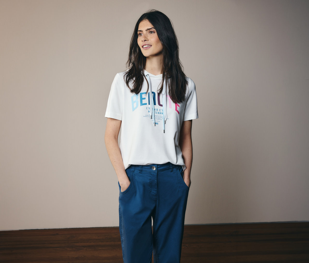 Nő kék nadrágban és fehér, nyomott mintás CECIL kapucnis pólóban, kezével a zsebében.