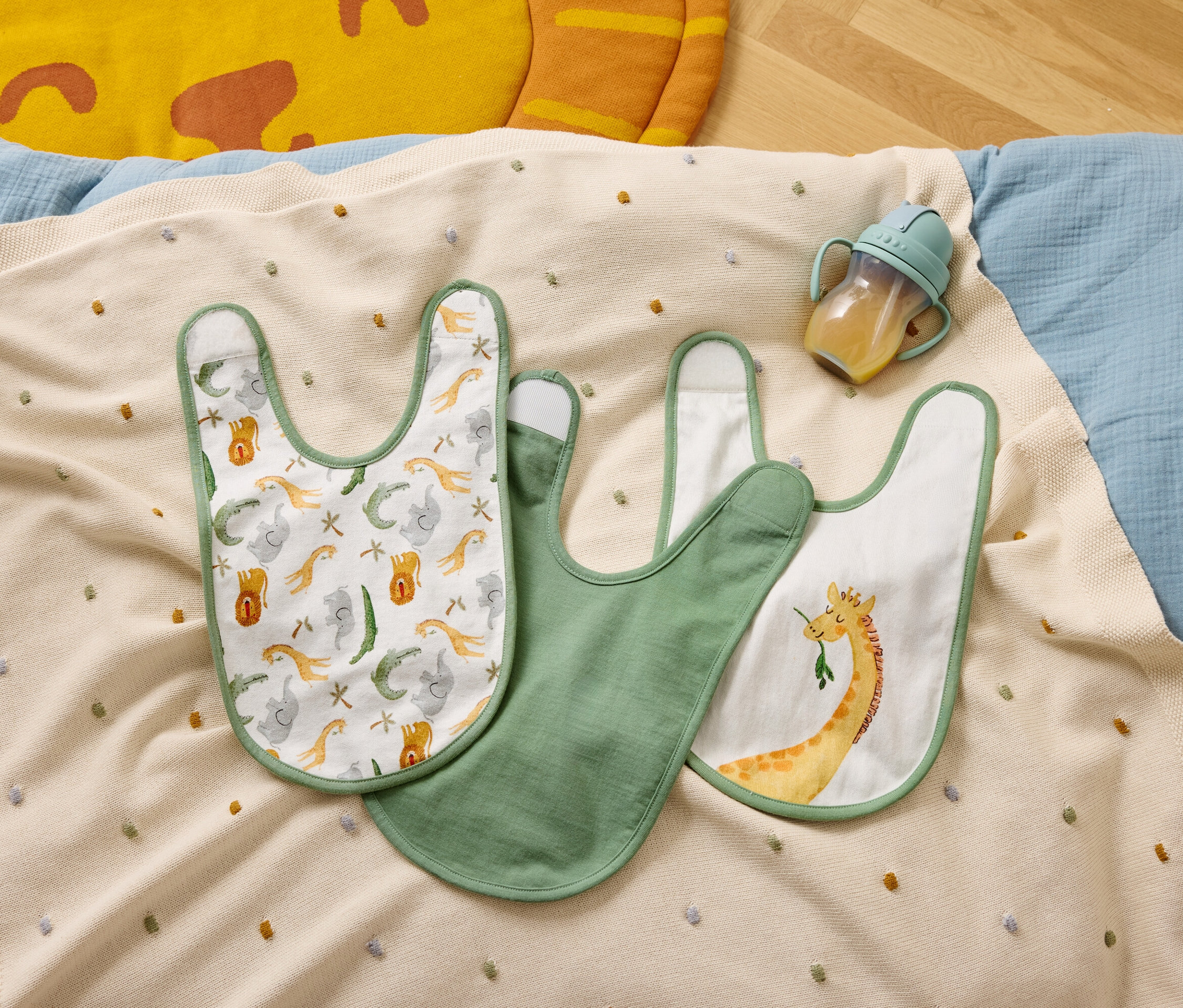 Három előke és egy cumisüveg fekszik egy bézs színű, színes pöttyös takarón.