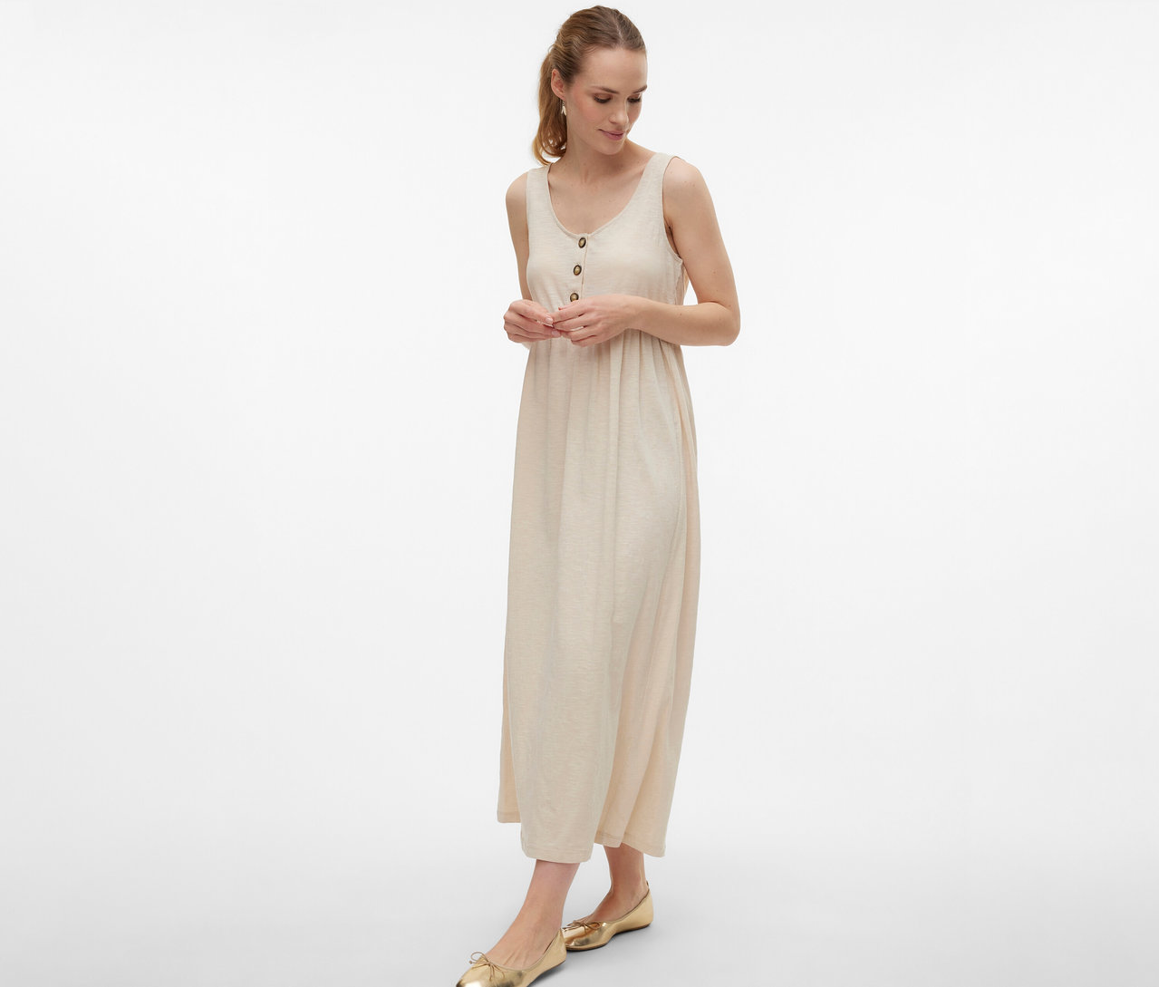 Nő visel gombos MAMALICIOUS »MLEVI LIA« kismamaruhát és arany balerinacipőt fehér háttér előtt.