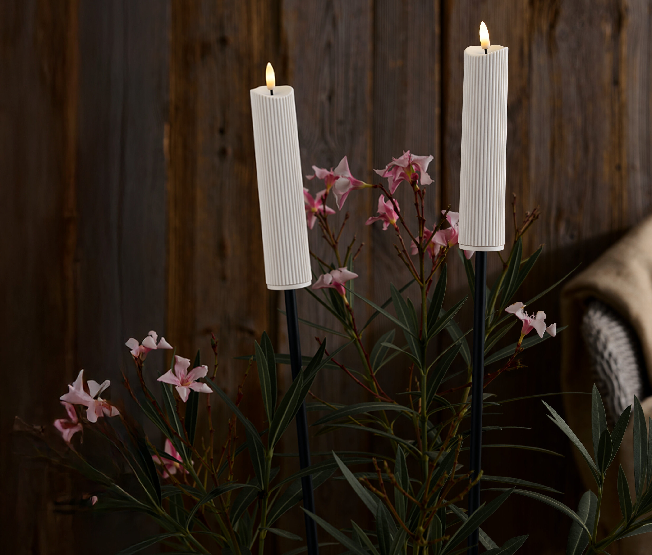 Két fehér, bordázott gyertya fekete állványokon, rózsaszín virágok és zöld levelek között, sötét fa háttér előtt.