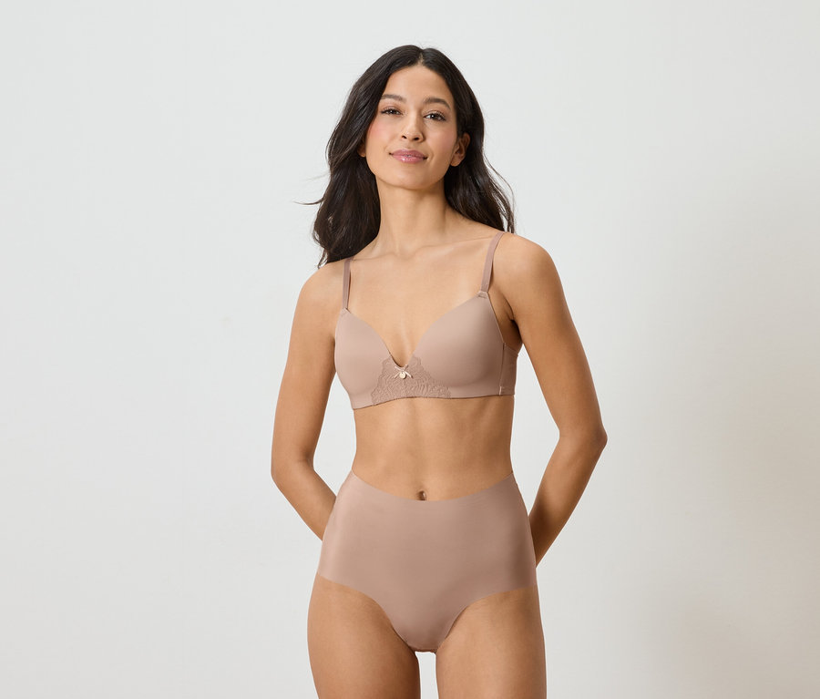 Nő visel testszínű melltartót és alsóneműt fehér háttér előtt.
