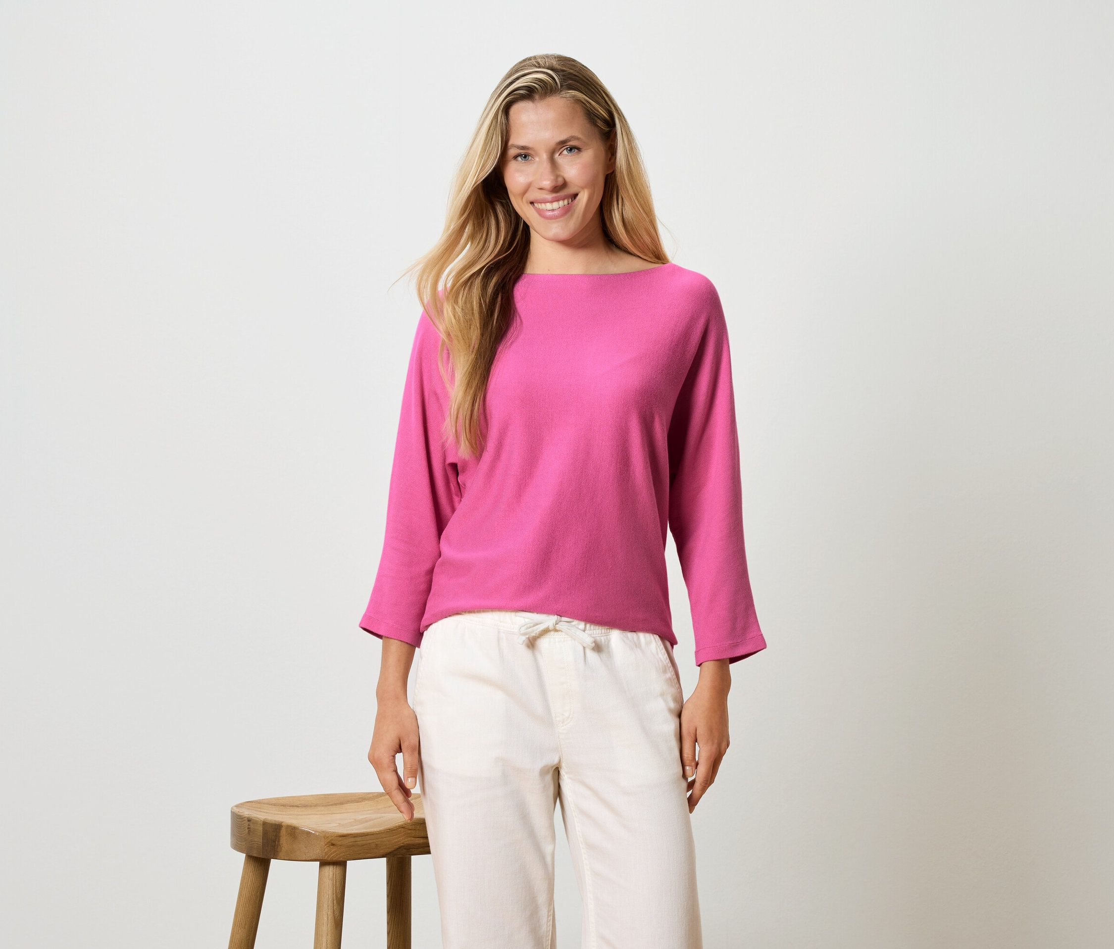 Egy hosszú szőke hajú nő rózsaszín, hosszú ujjú felsőt és világos nadrágot visel, egy zsámoly mellett áll.