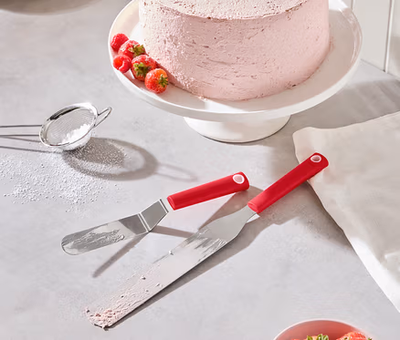 Egy rózsaszín torta eperrel és málnával egy fehér tortatálon áll, mellette két piros spatula, egy tál eper és egy porcukorszita.