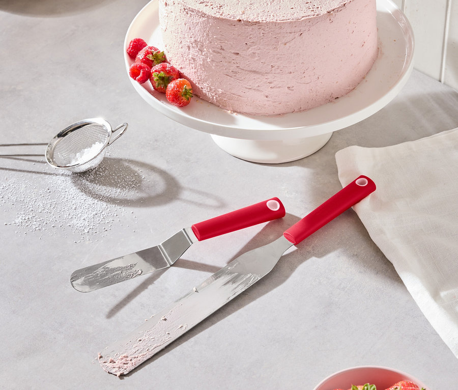 Egy rózsaszín torta eperrel és málnával egy fehér tortatálon áll, mellette két piros spatula, egy tál eper és egy porcukorszita.