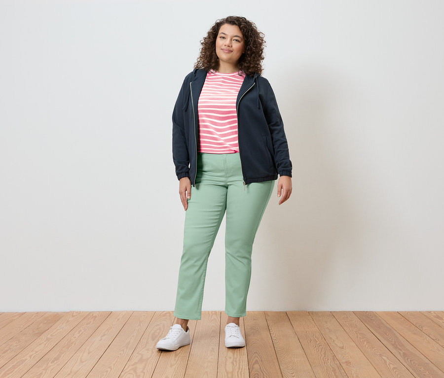 Egy nő áll egy világos környezetben, sötétkék kabátot, csíkos felsőt, világoszöld nadrágot és fehér tornacipőt visel.