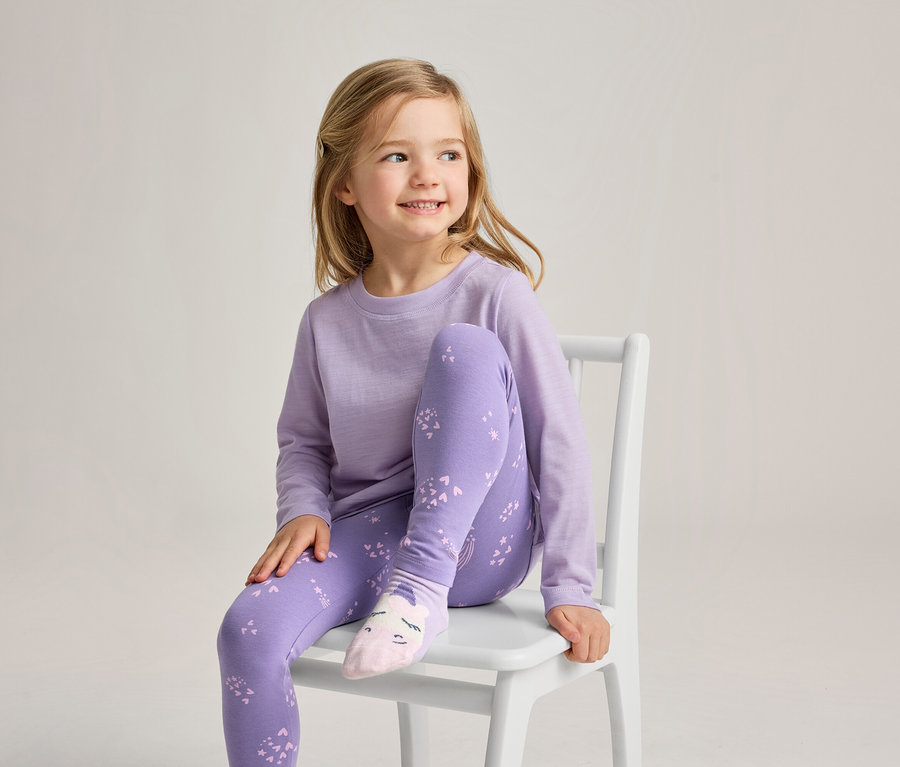 Egy fiatal szőke hajú lány ül egy fehér széken, mosolyogva jobbra néz. Világoslila hosszú ujjú felsőt, lila leggingst világosabb lila mintával és világos rózsaszín zoknit visel, amelyen egy arc van.
