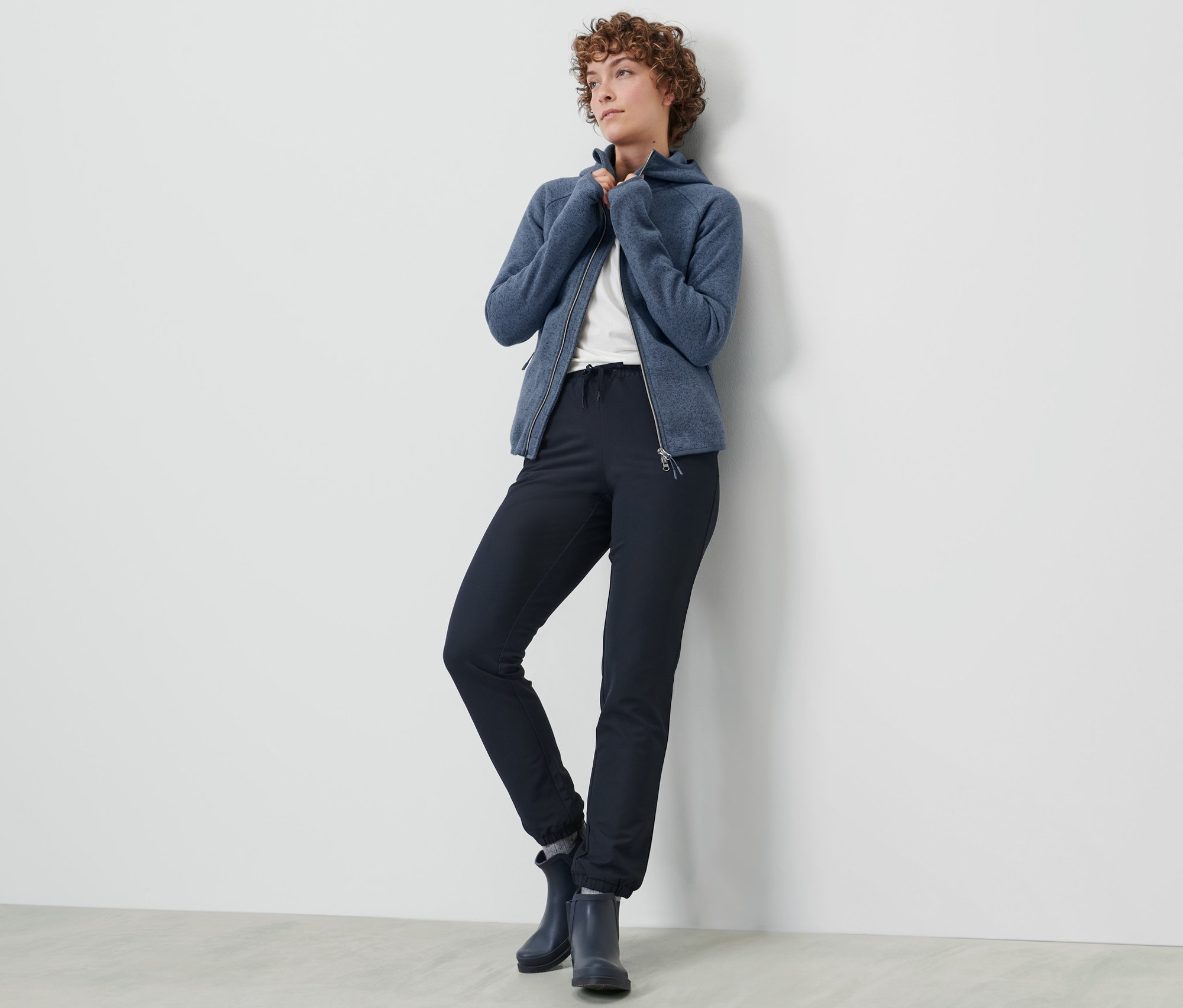 Nő pózol softshell szabadidőnadrágban és kék cipzáras pulóverben a falnál.