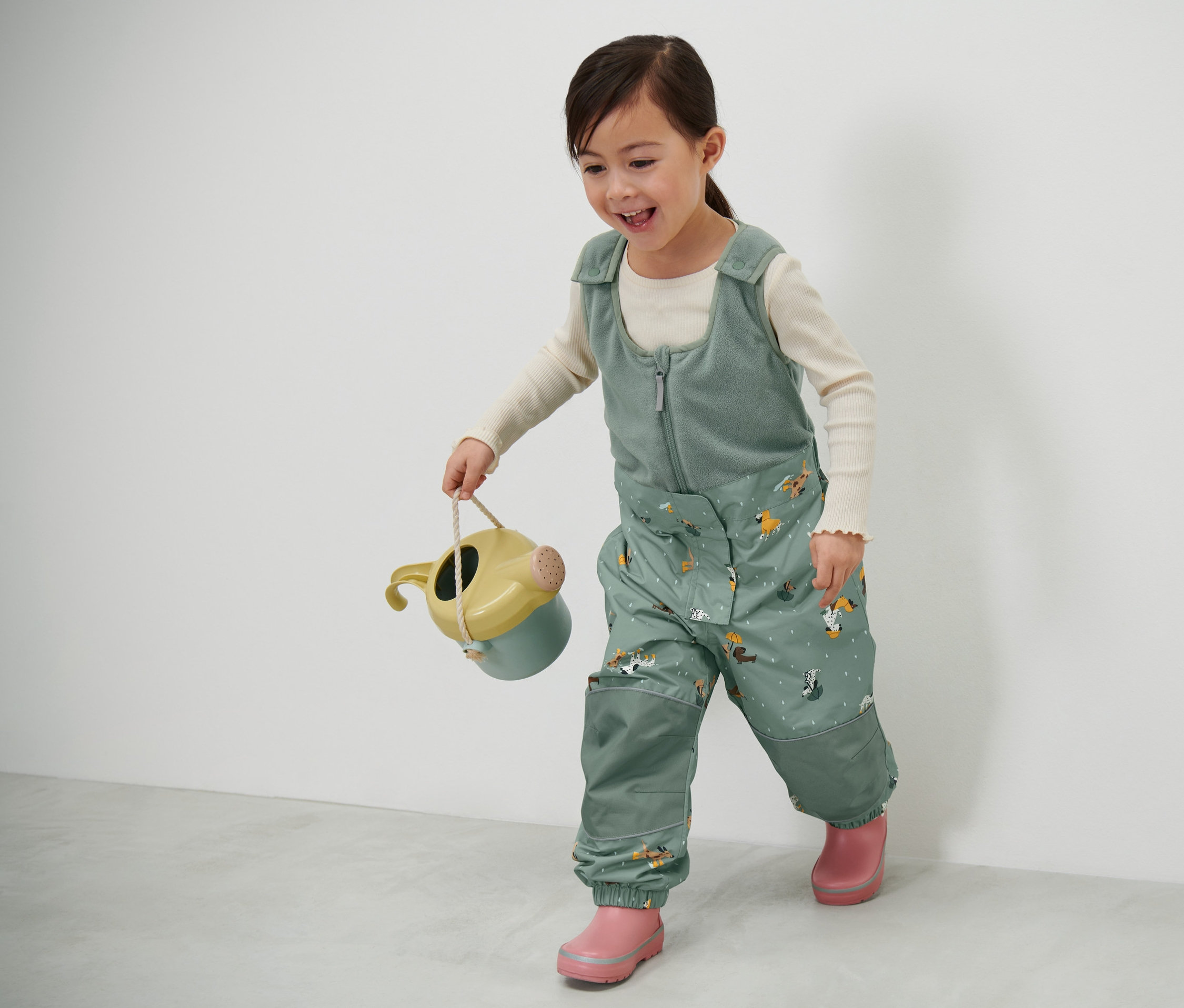 Kislány gyerek termo esőnadrágot visel, kutyás mintával, és egy locsolókannát tart.