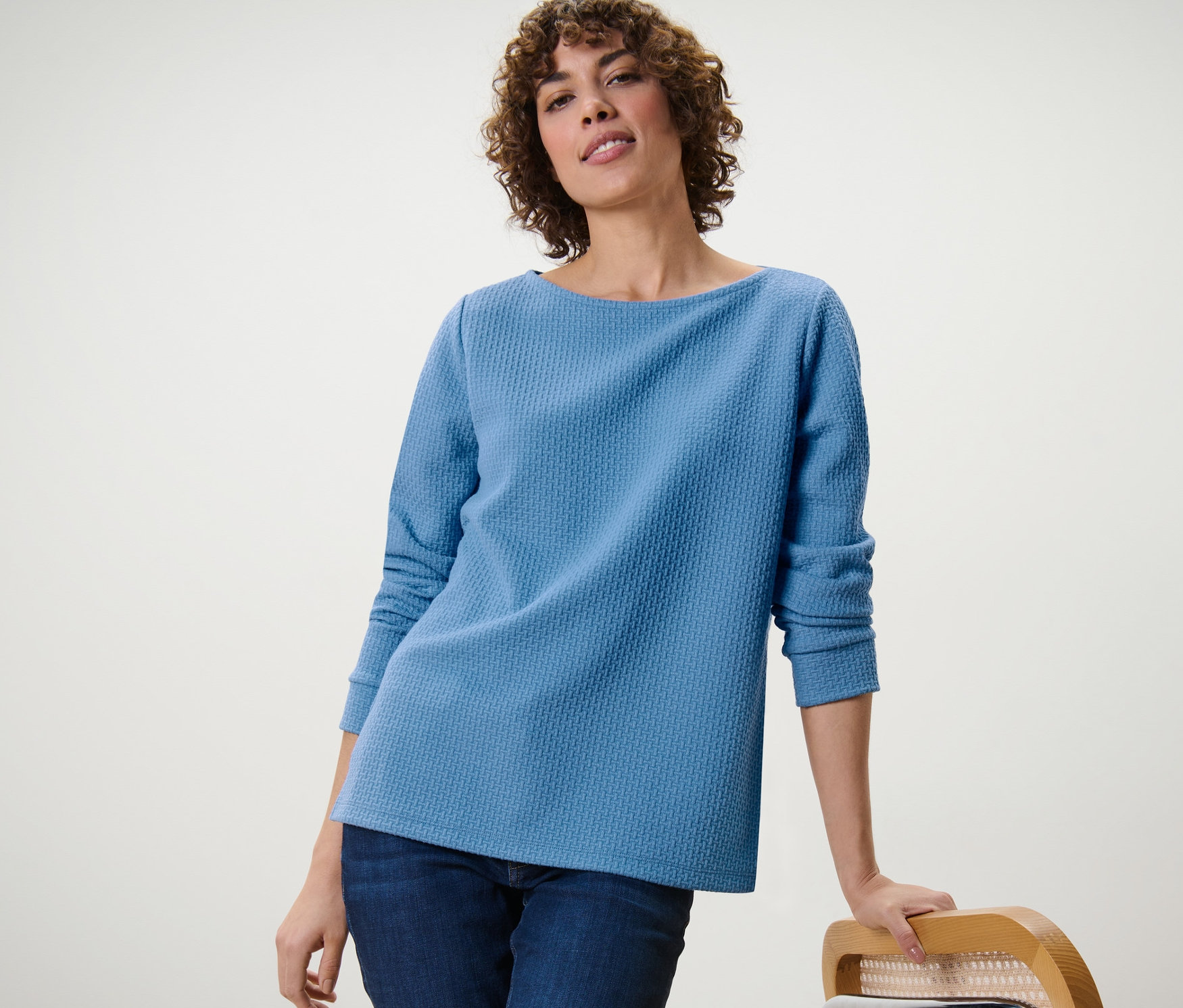 Egy nő egy széknek támaszkodva világoskék, strukturált női szabadidőfelsőt visel.