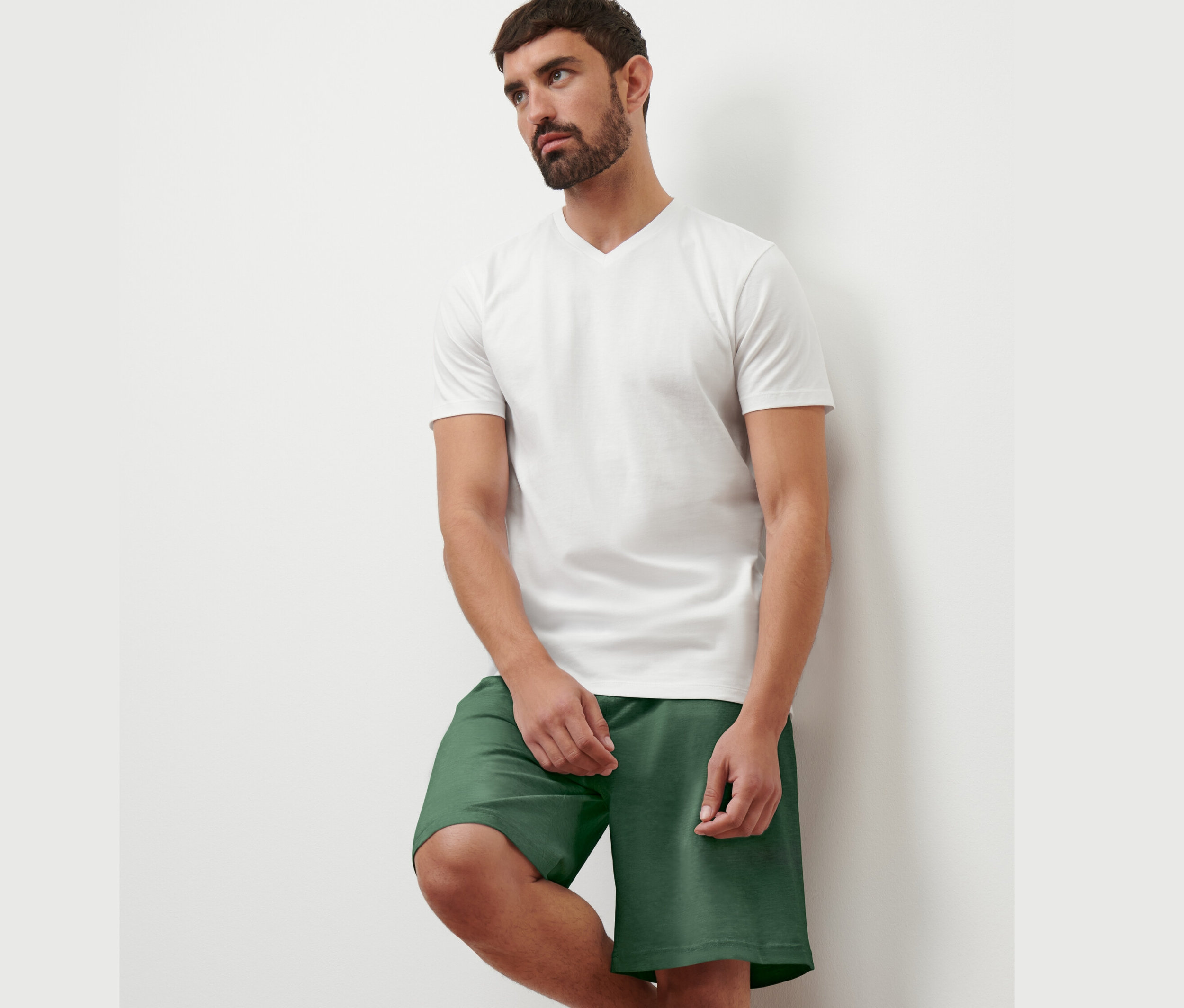 Férfi támaszkodik a falnak, fehér V-nyakú pólót és zöld rövidnadrágot visel. A 2 férfi V-nyakú pólóból egyet visel.