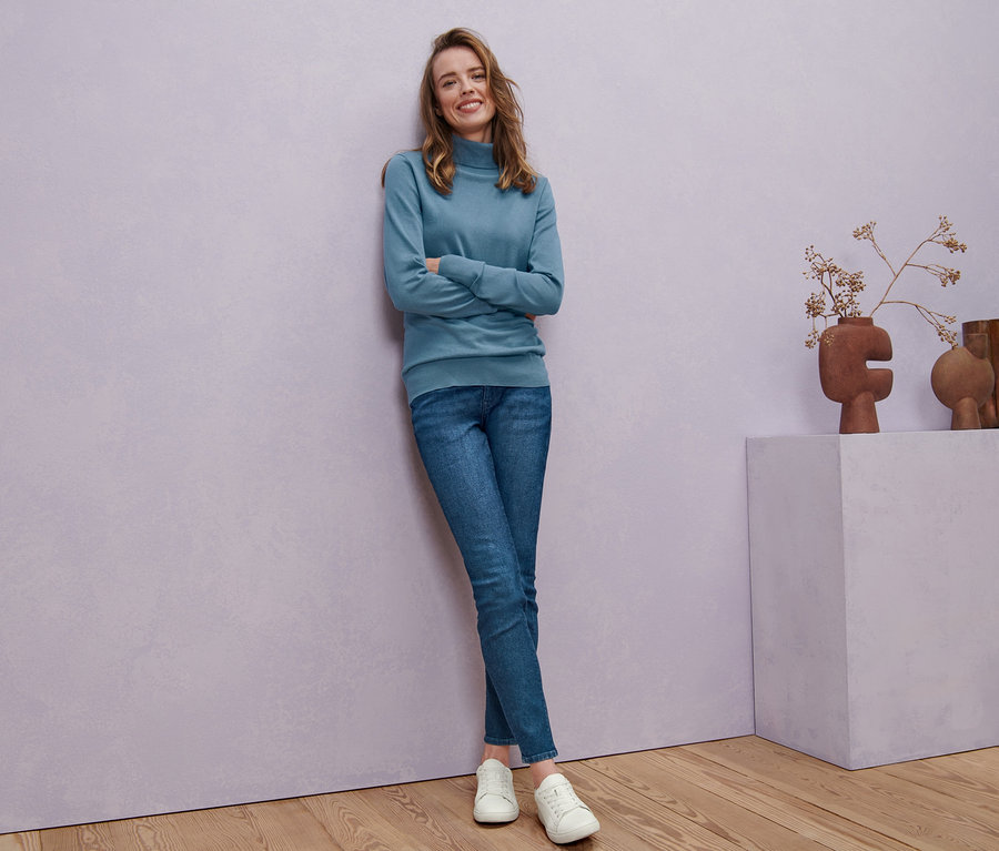 Nő pózol füstkék, finom kötésű garbónyakú pulóverben, kék skinny farmernadrágban és fehér bőr sneaker cipőben.
