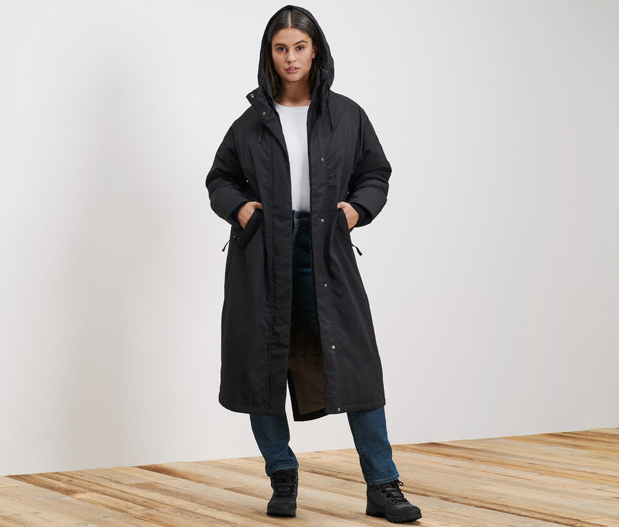 Egy nő áll, és extra hosszú termokabátot és sötétkék Slim Fit Emma farmernadrágot visel.