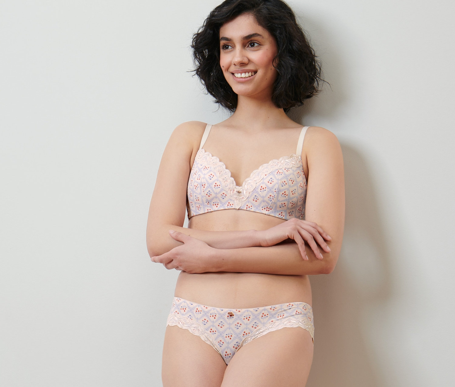 Mosolygó nő pózol krémszínű, nyomott mintás csipkés fehérnemű szettben. 2 női brazil alsót visel.