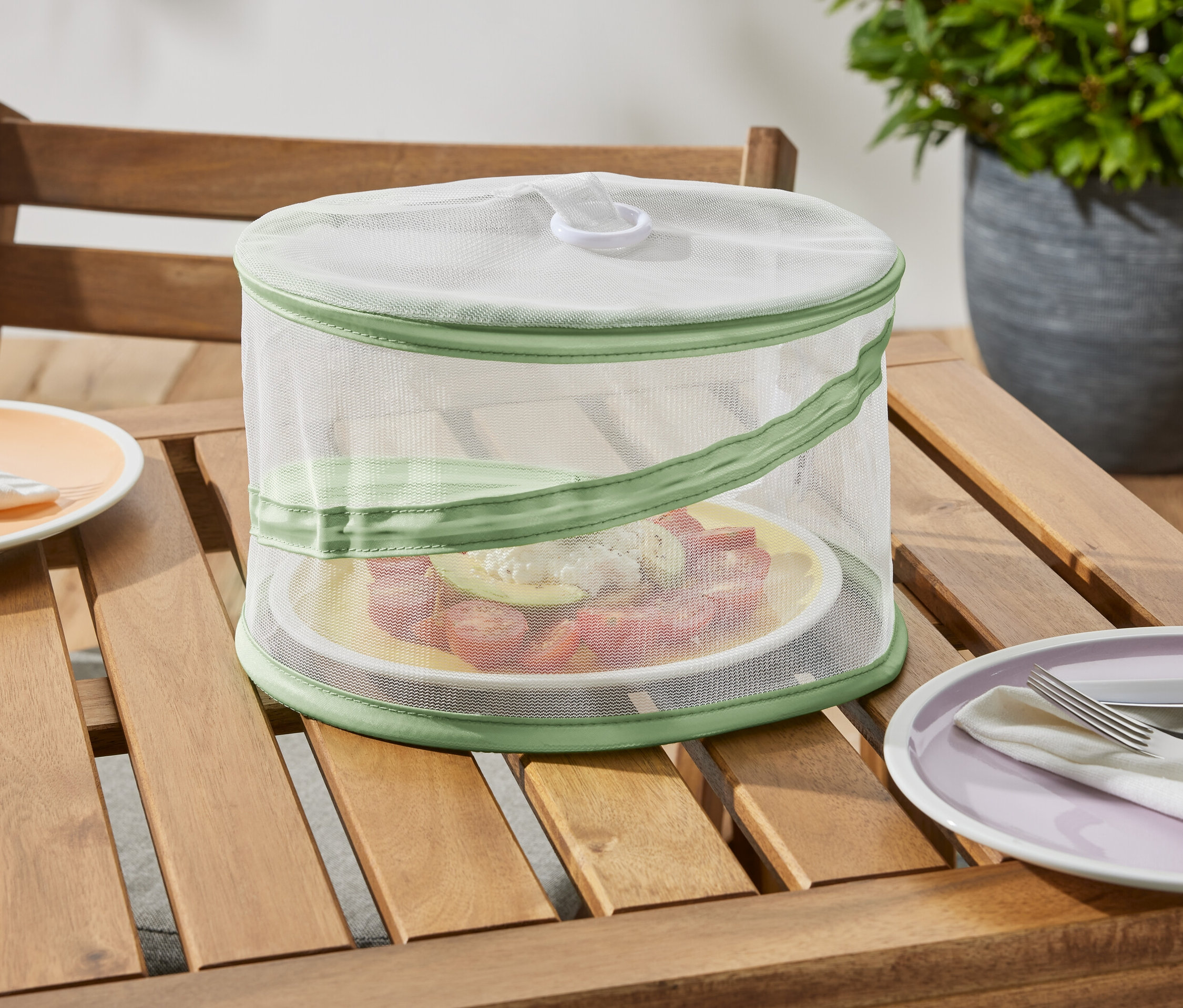 Az Összehajtható ételfedő háló, mini egy fa asztalon áll, és megvéd egy tányér ételt.