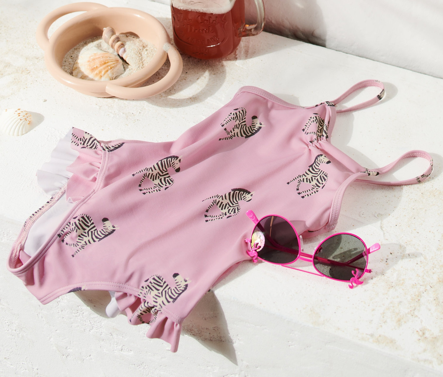 Egy rózsaszín, zebrás mintázatú, fodros fürdőruha egy rózsaszín napszemüveg, egy üdítős pohár és egy strandtáska mellett fekszik.