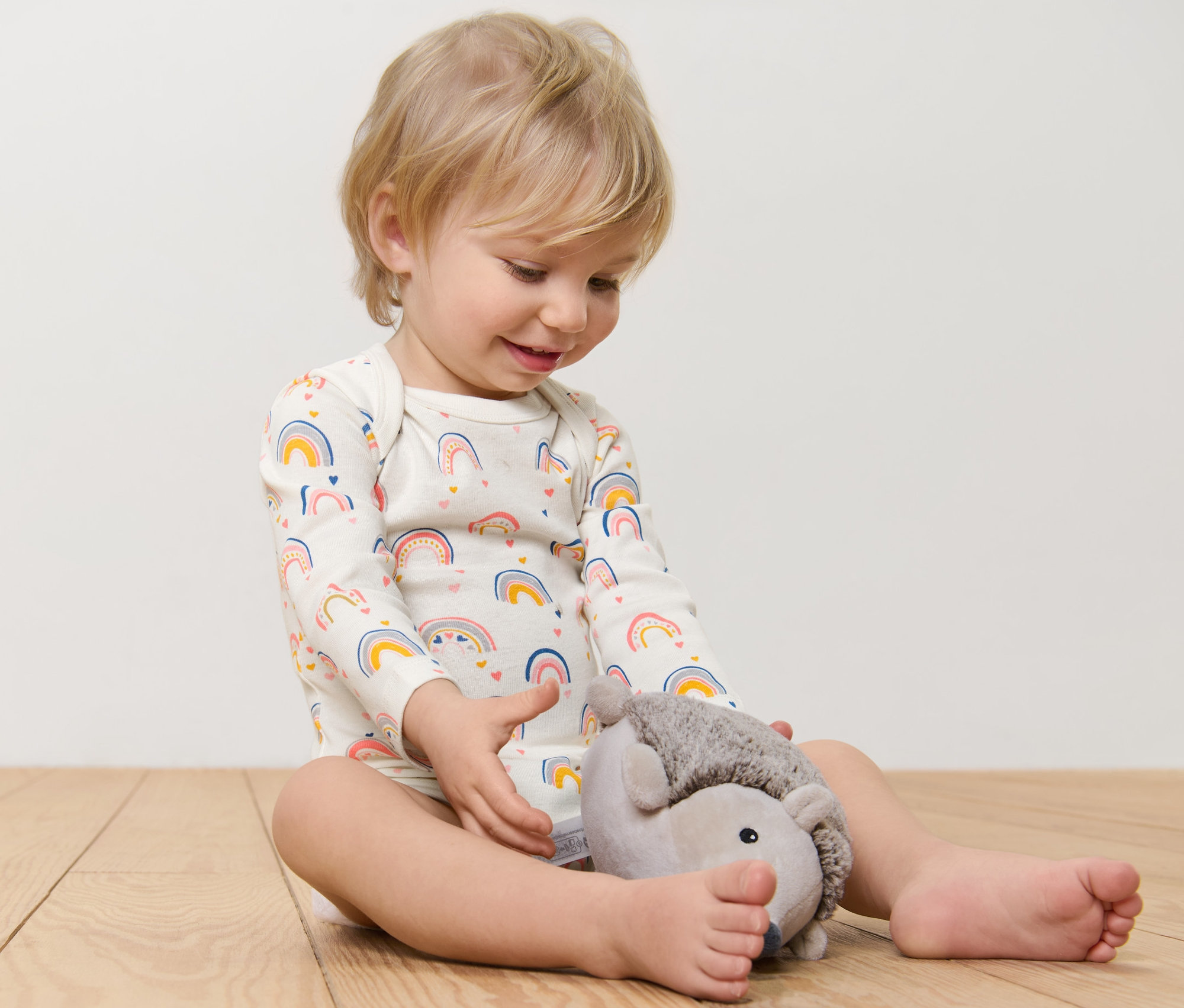 Egy gyermek ül a padlón, és egy plüss sünivel játszik. Szivárvány mintás, hosszú ujjú bodyt visel.