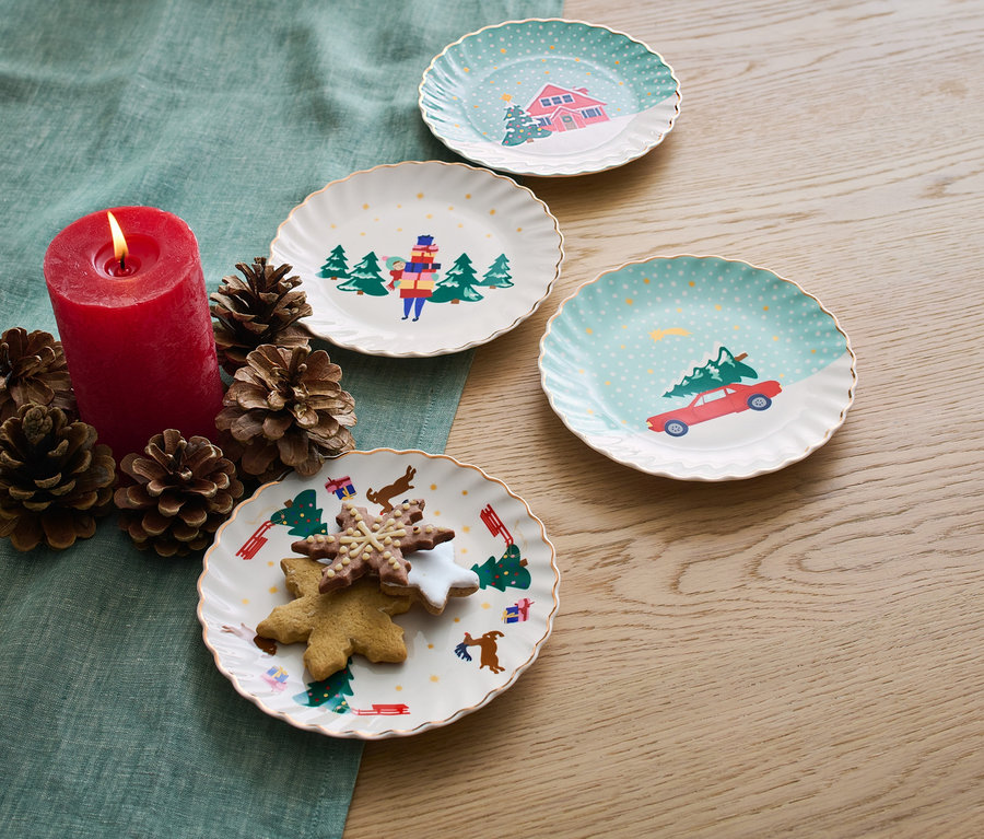 Négy karácsonyi desszertes tányér, gyertya, fenyőtobozok és sütemények az asztalon.