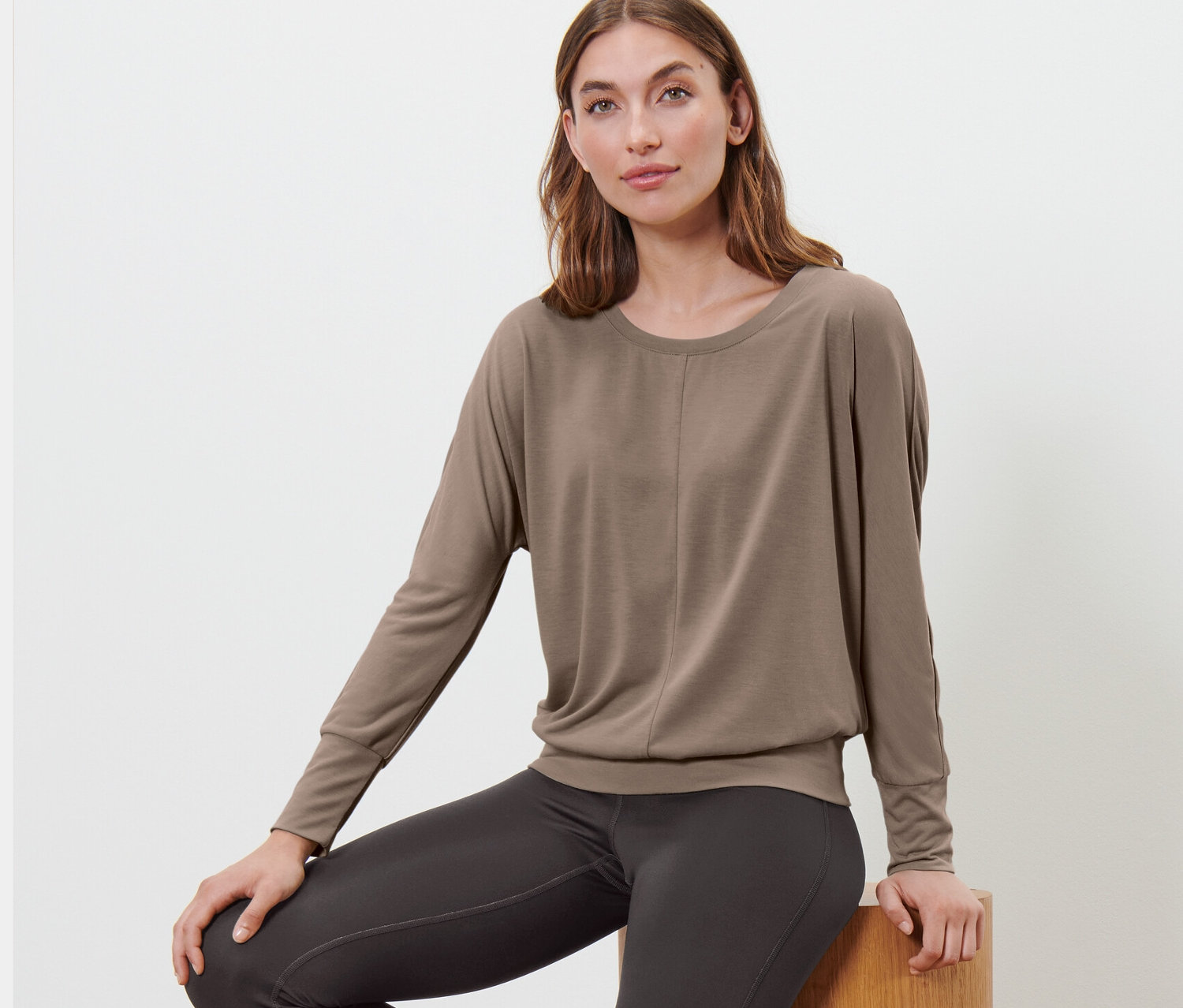 Nő ül fa lépcsőn, és taupe színű női denevérujjú sportfelsőt visel.