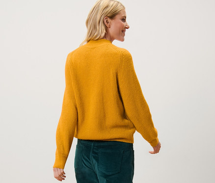 Nő sétál egy mustársárga laza kötésű pulóverben és zöld nadrágban.