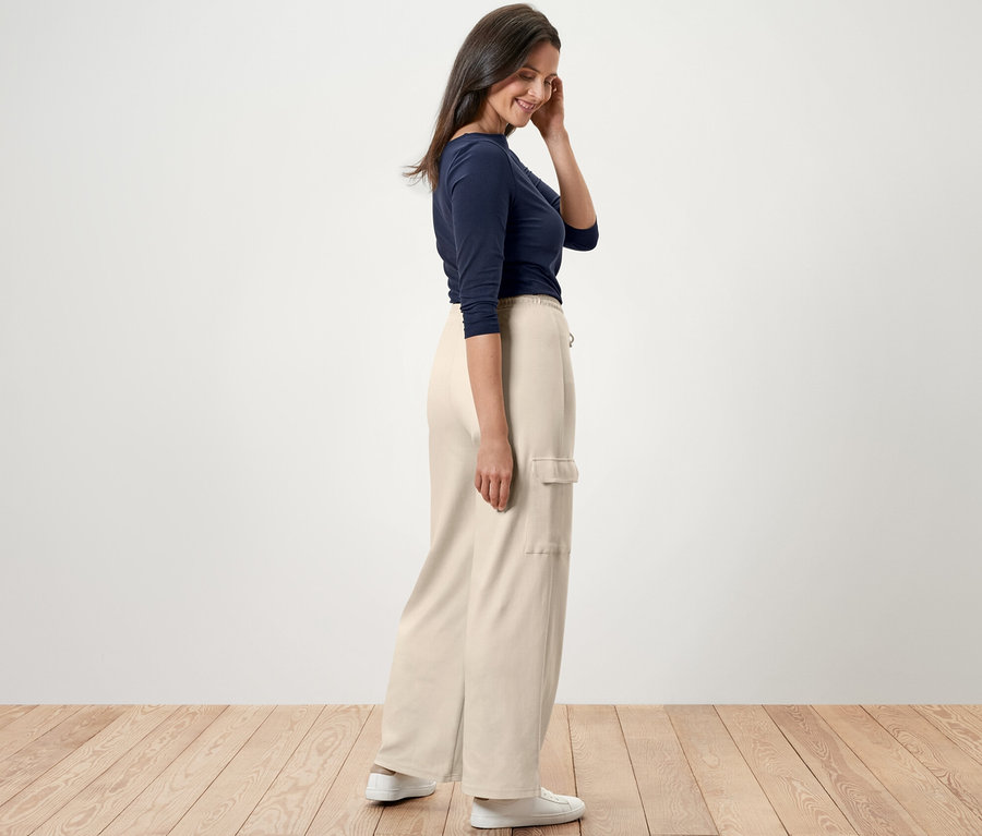 Egy nő oldalra állva krémfehér szabadidőnadrágot, kék pólót és fehér tornacipőt visel.