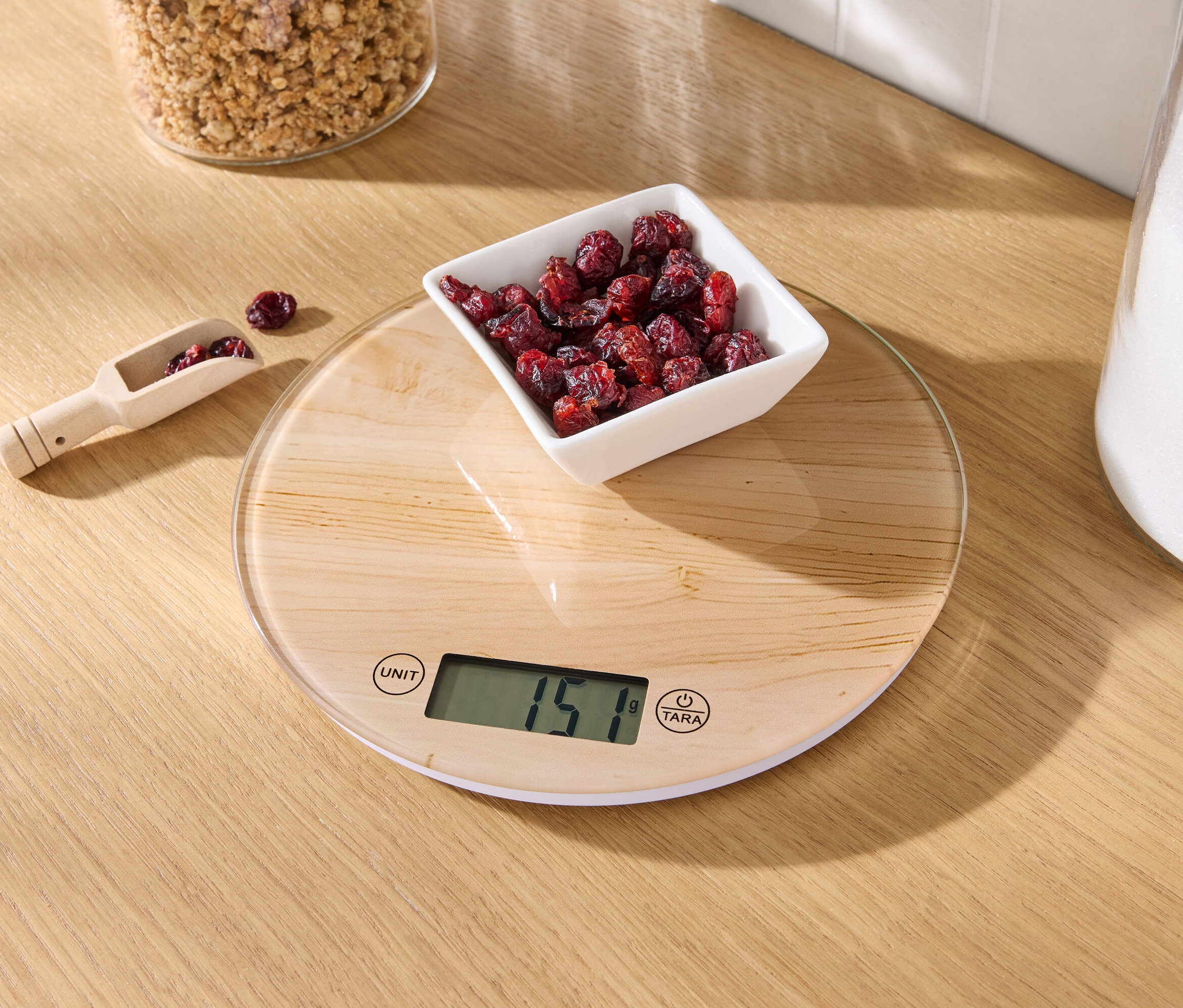 A fa asztalon egy konyhai mérleg látható egy tál szárított áfonyával és egy fából készült mérőkanál áfonyával.