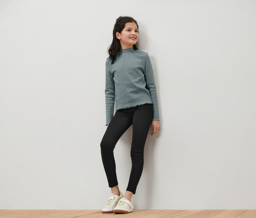 Mosolygó tinédzser lány kékeszöld, hosszú ujjú felsőben, fekete farmerben és fehér tornacipőben pózol egy fehér fal előtt.