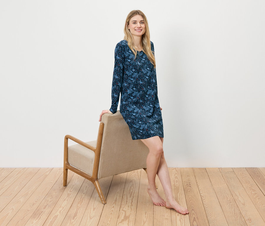 Egy nő egy bézs színű széknek dőlve ül, kék virágos ruhát visel.