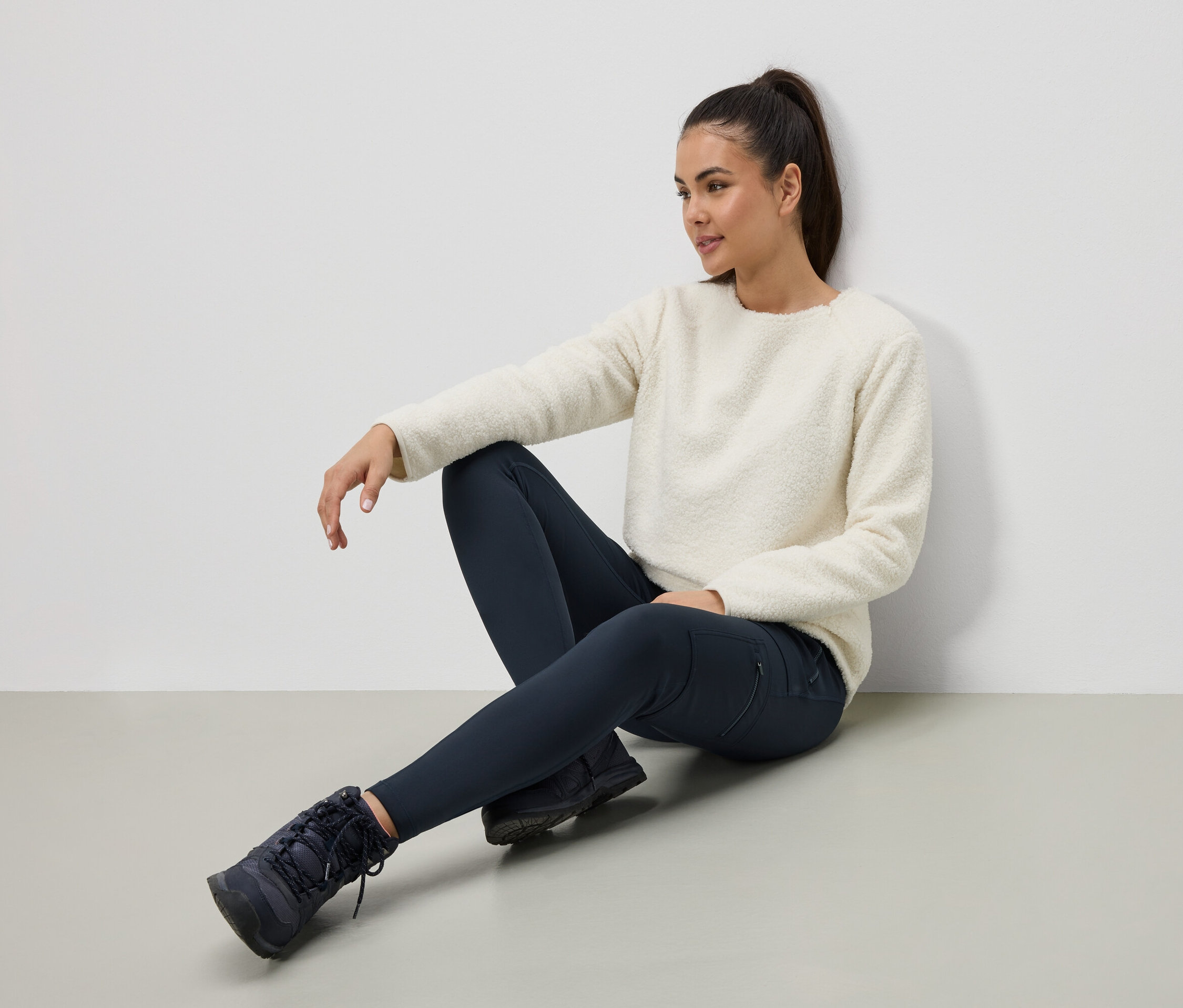 Nő ül a padlón krémszínű pulóverben, sötétkék leggingsben és csizmában.