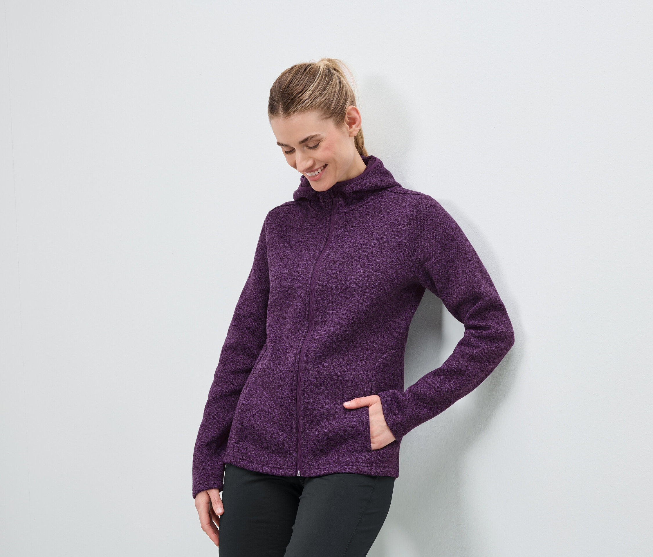 Egy nő lila, cipzáros kapucnis pulóverben és fekete nadrágban áll egy fehér fal mellett.