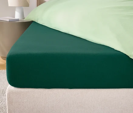Egy zöld gumis lepedő közeli képe egy ágyon, krémszínű fejtámlával és világoszöld párnahuzattal.