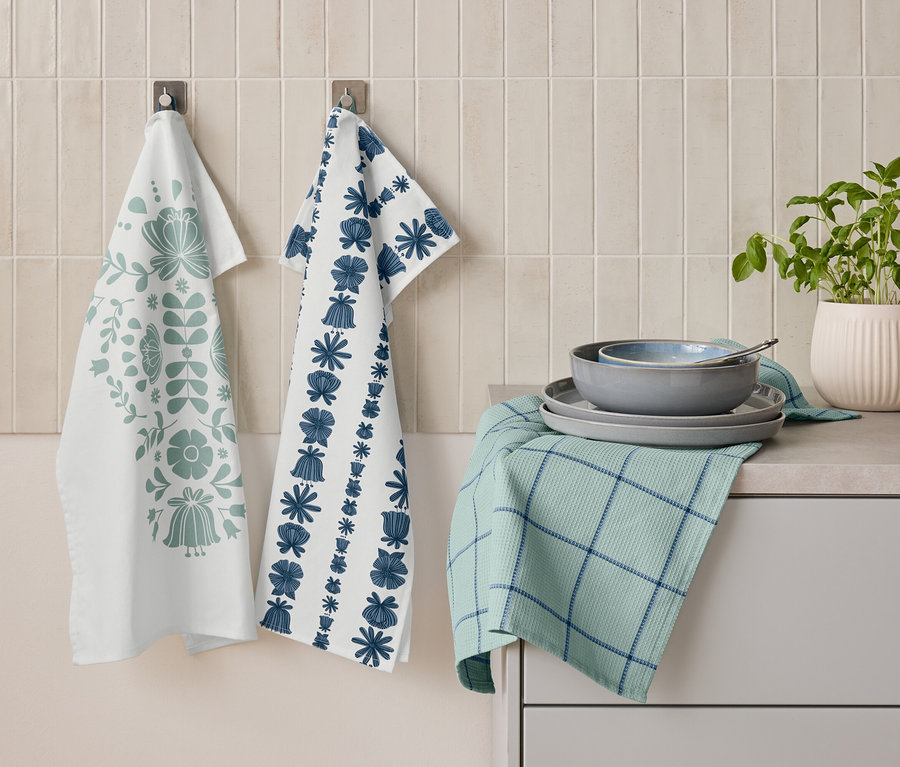 Két virágmintás konyharuha lóg egymás mellett a falon. Alattuk egy összehajtott zöld, kék csíkos konyharuha, rajta tányérok és egy tál egymásra rakva. Mellette egy cserép növény áll.