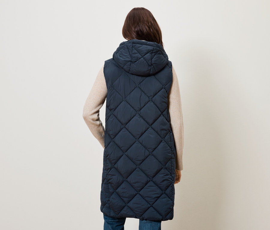 Hátulról látható nő hosszú, steppelt, sötétkék mellényt visel kapucnival.