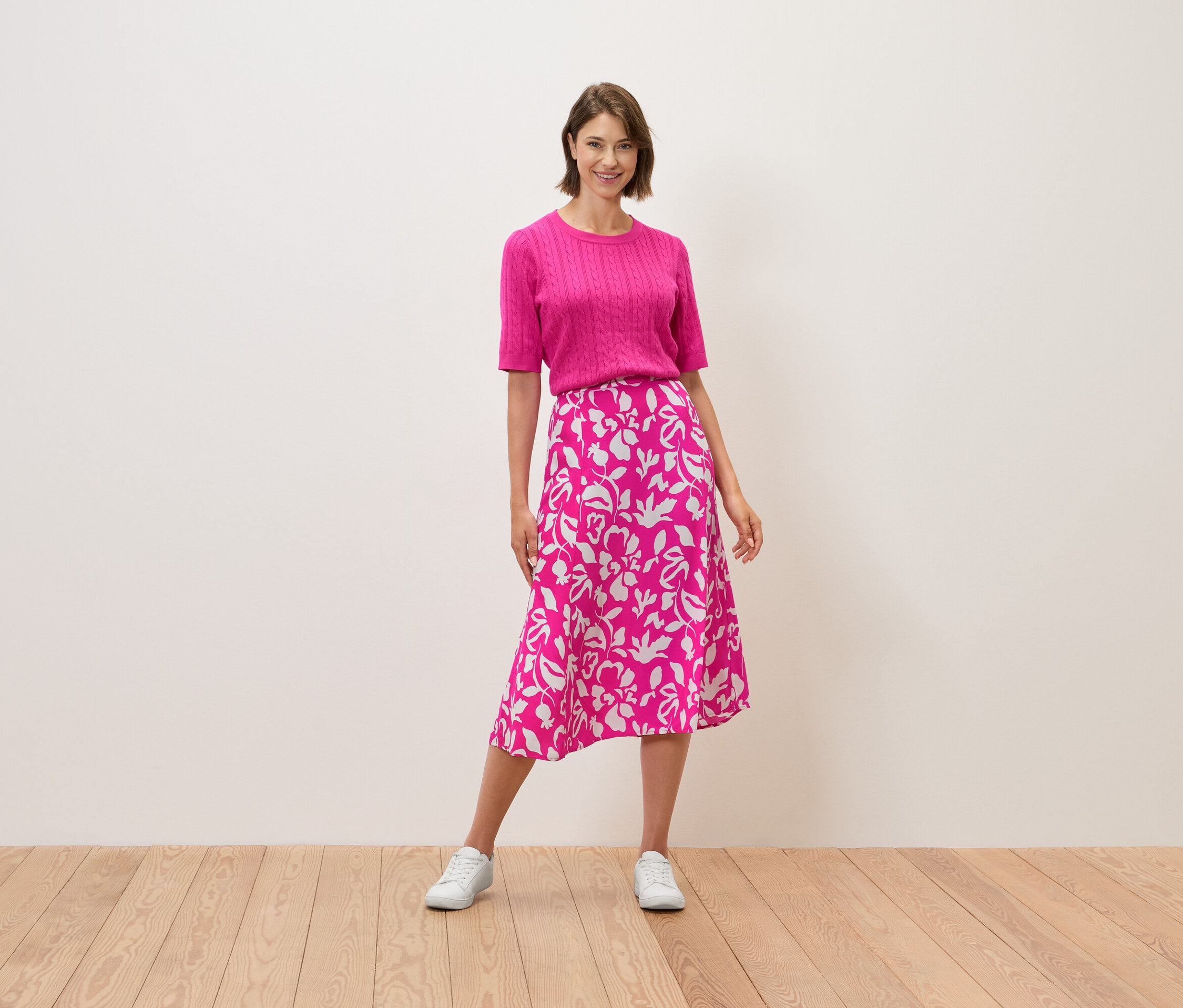 Egy nő áll, rózsaszín felsőt és virágmintás szoknyát visel.