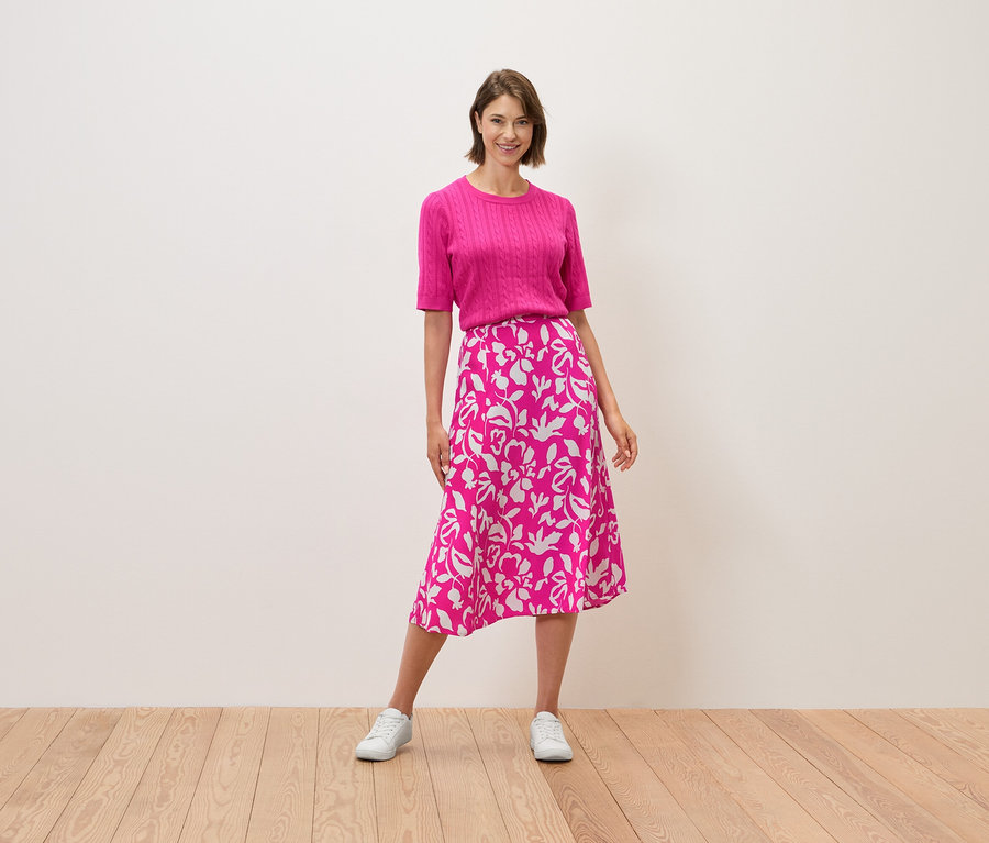 Egy nő áll, rózsaszín felsőt és virágmintás szoknyát visel.