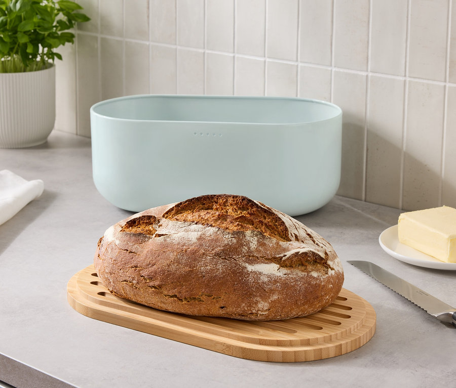 Egy kenyér egy fa vágódeszkán egy kék kenyértartó, egy vajdarab és egy kés mellett egy pulton.