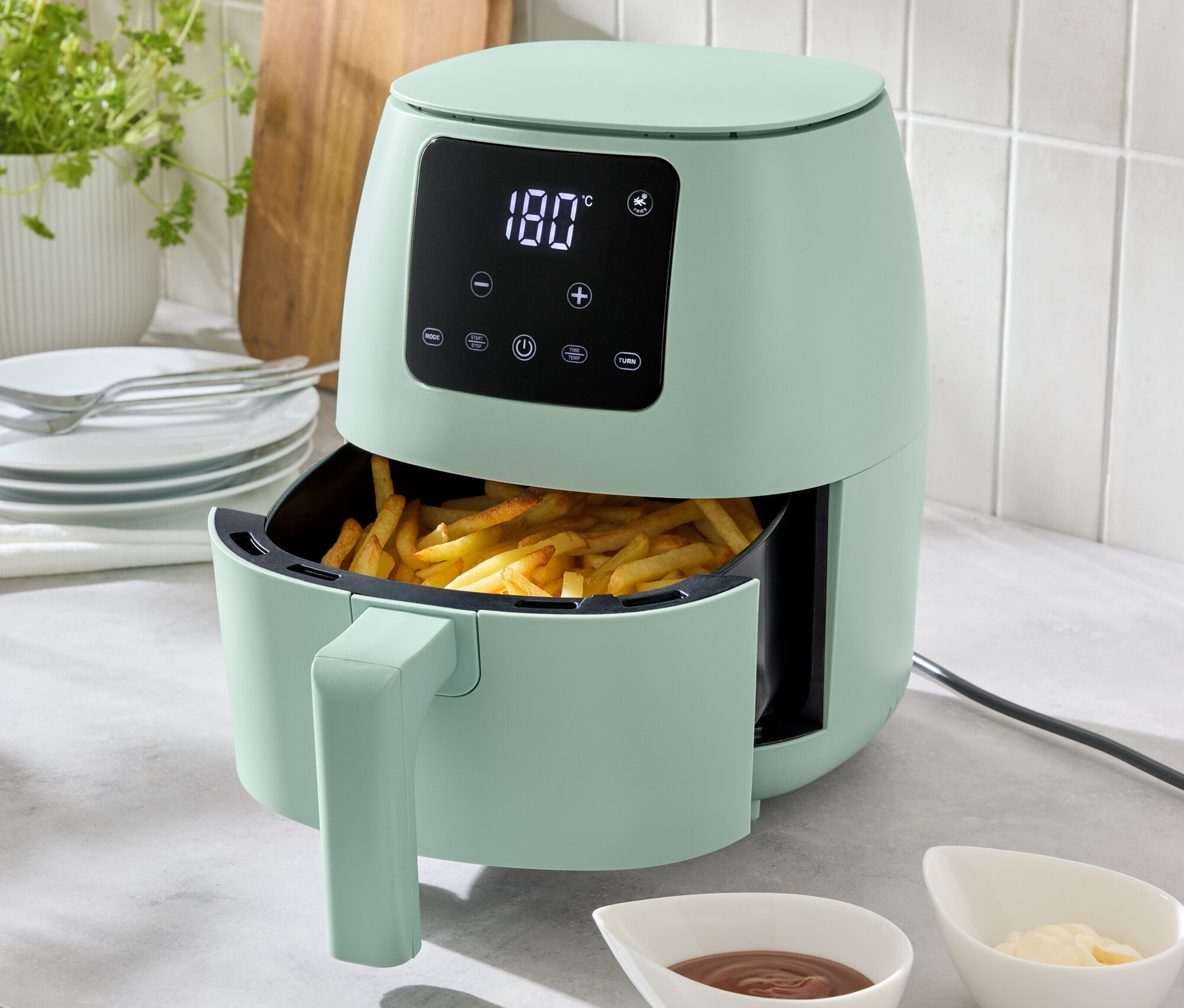 Egy világoszöld, 2 literes forrólevegős fritőz áll a konyhapulton, tele sült krumplival. Előtte két tálka mártás található.