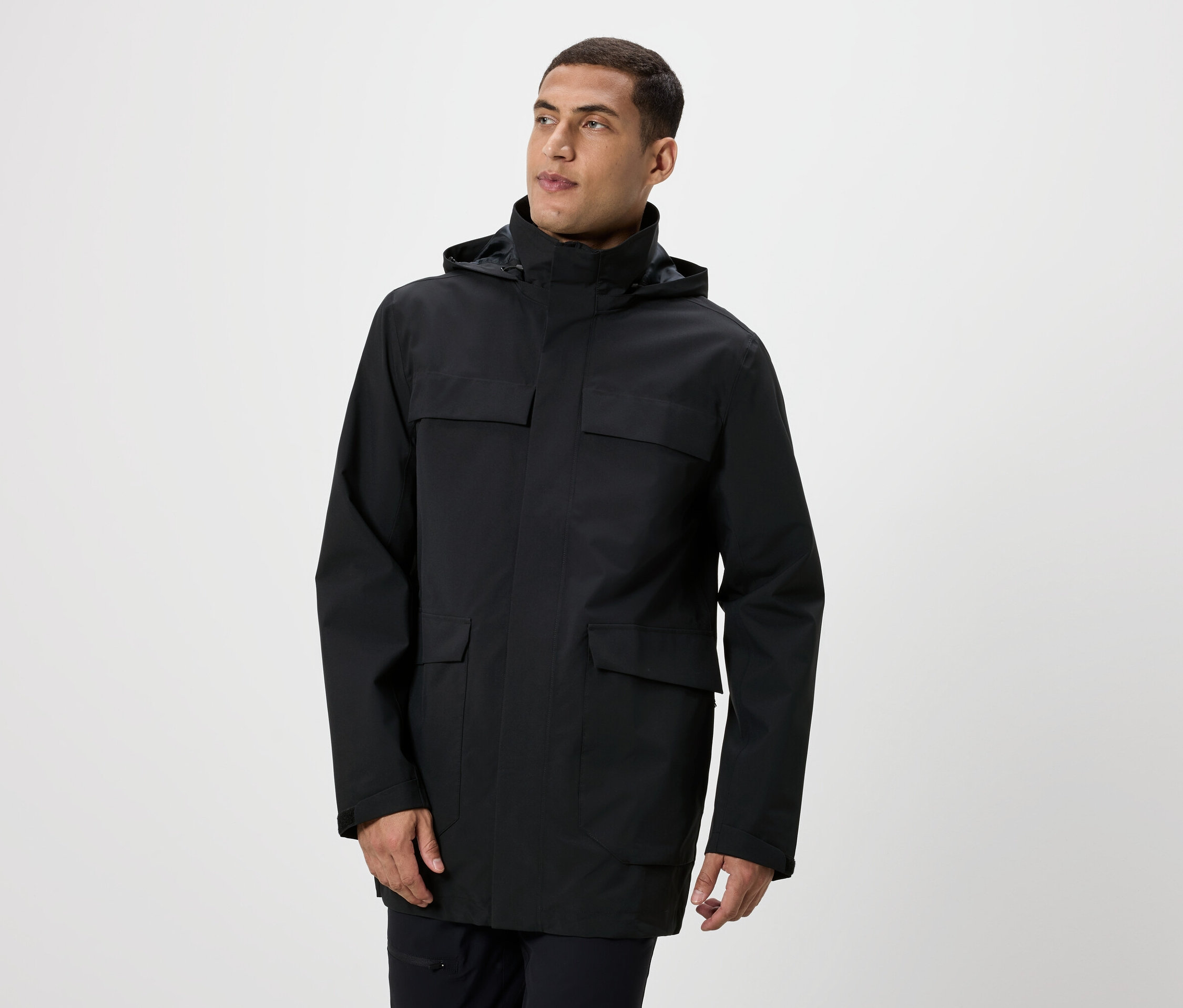 Férfi pózol fekete, kapucnis, zsebes kabátban.