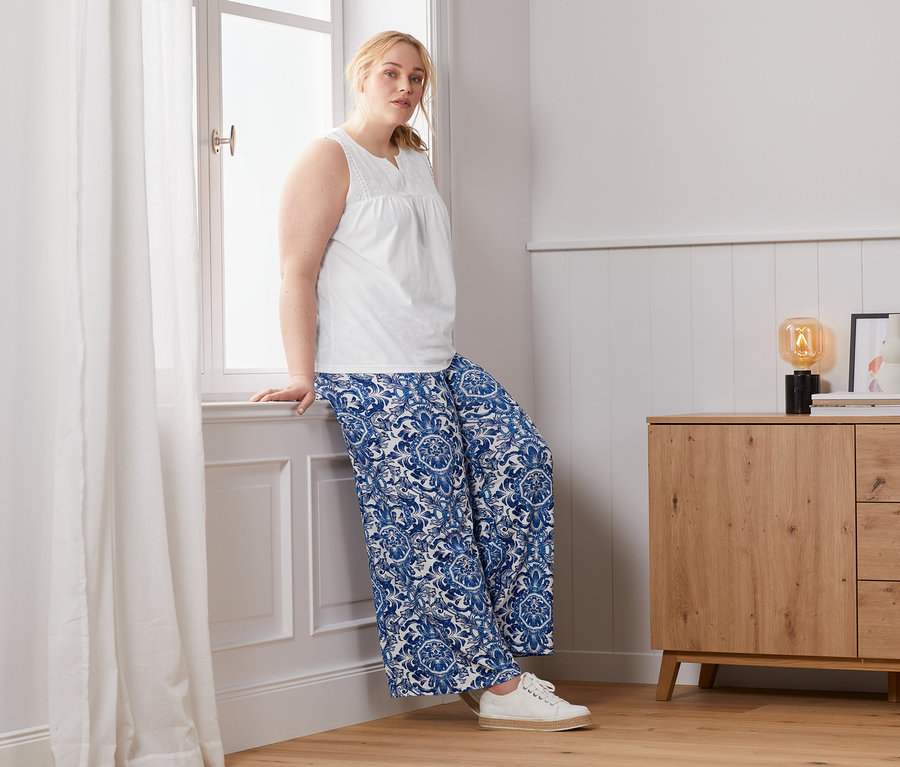 Nő támaszkodik az ablaknak hímzett toppban és mintás, bőszárú nadrágban.