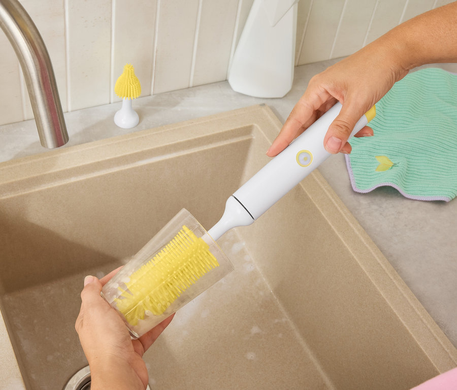 Egy személy egy sárga kefével tisztít egy poharat egy bézs mosogatóban. A háttérben egy fehér flakon és egy zöld törlőkendő látható.