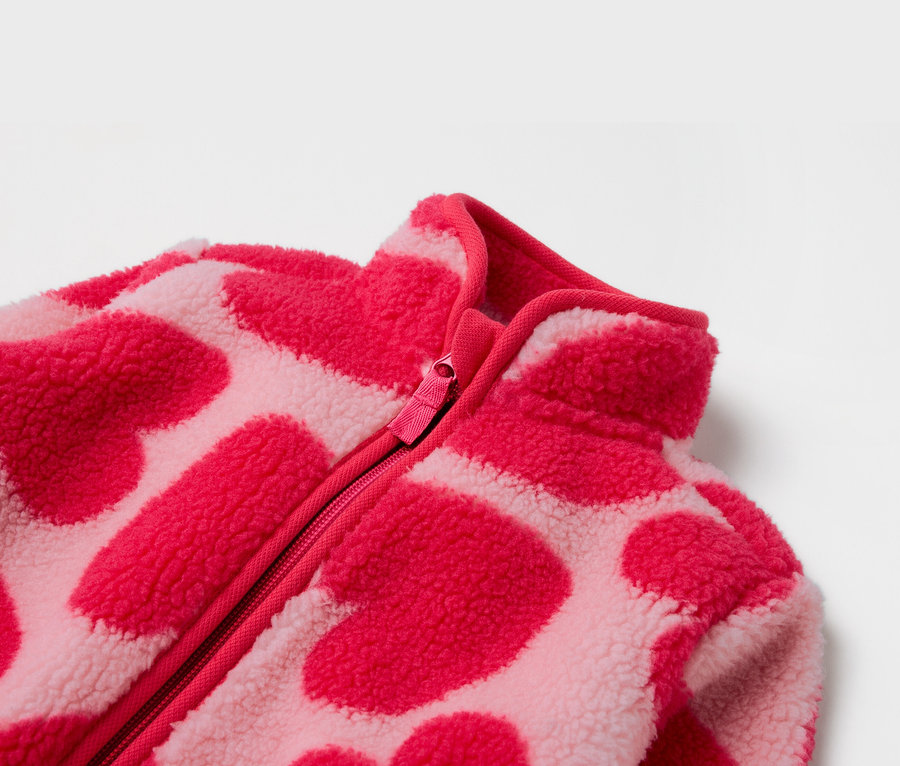 Egy rózsaszín, cipzáras, szívecskés mintázatú fleece kabát részlete.
