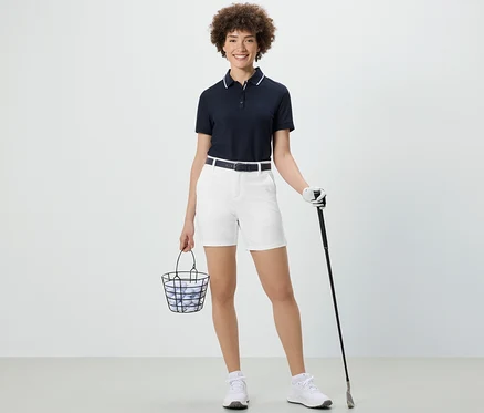 Nő tart egy kosár golf labdát és egy golfütőt. Fehér funkcionális softshell rövidnadrágot visel.