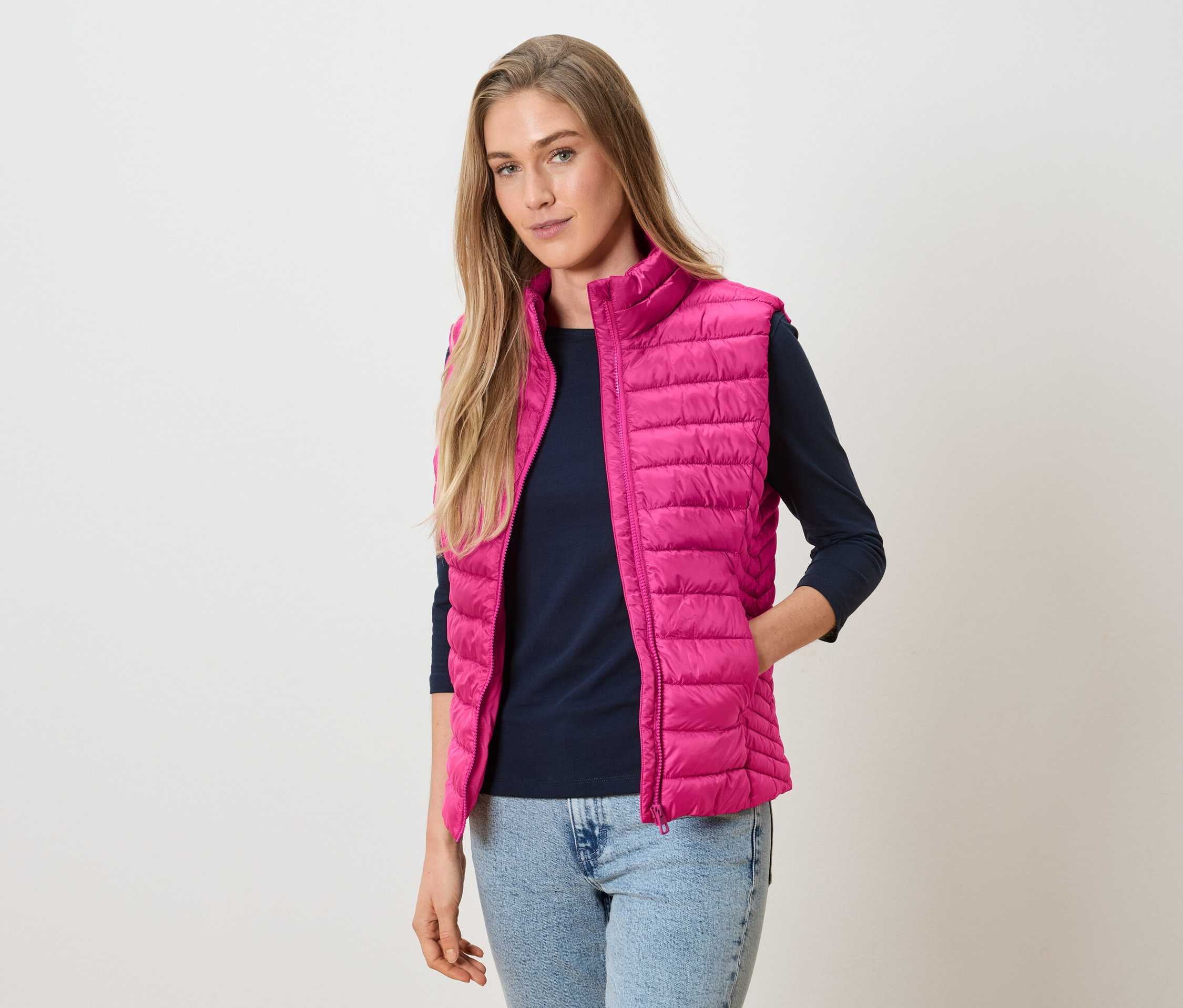 Nő visel rózsaszín steppelt mellényt egy tengerészkék hosszú ujjú felső felett, farmerrel.